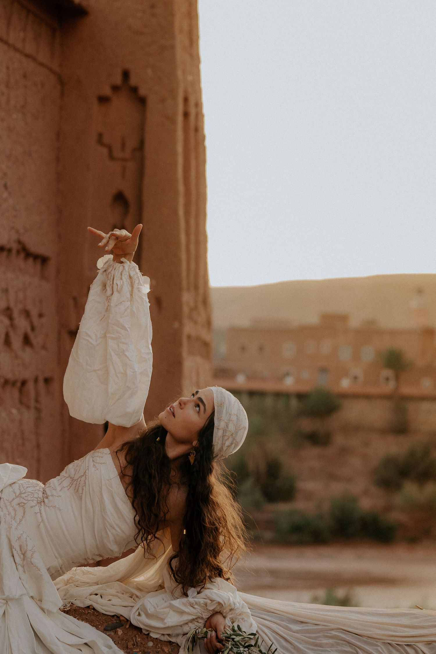 Woman in flowing white dress poses gracefully against desert architecture at golden hour.