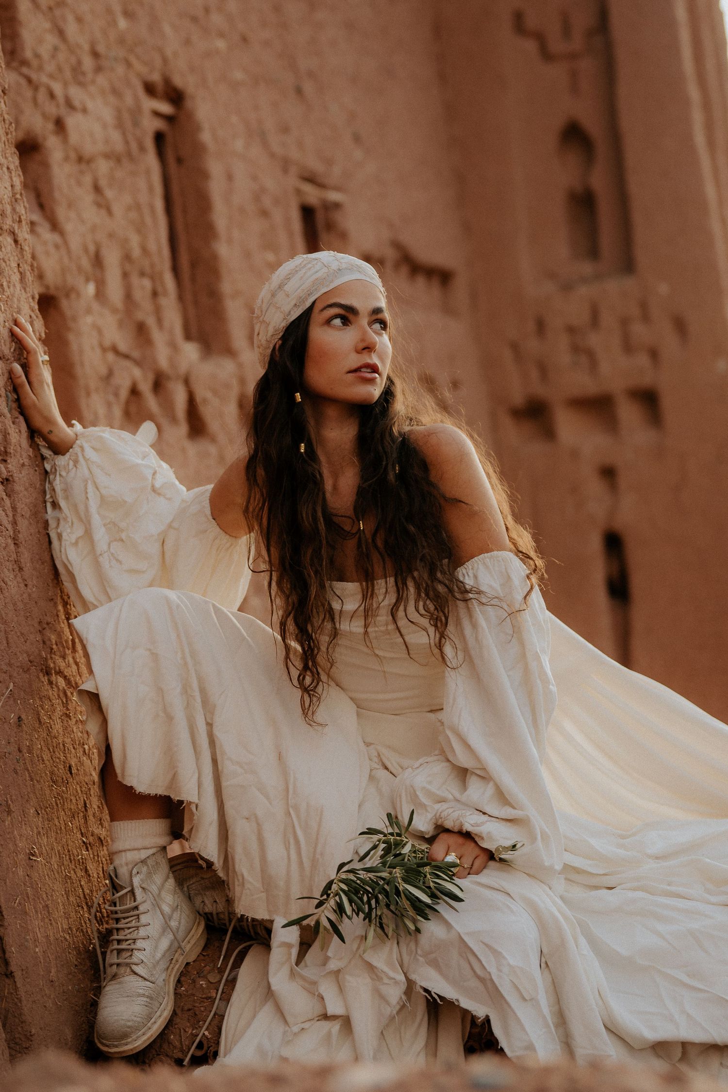 A person in a flowing white dress and sneakers poses against a textured adobe wall in warm sepia tones.
