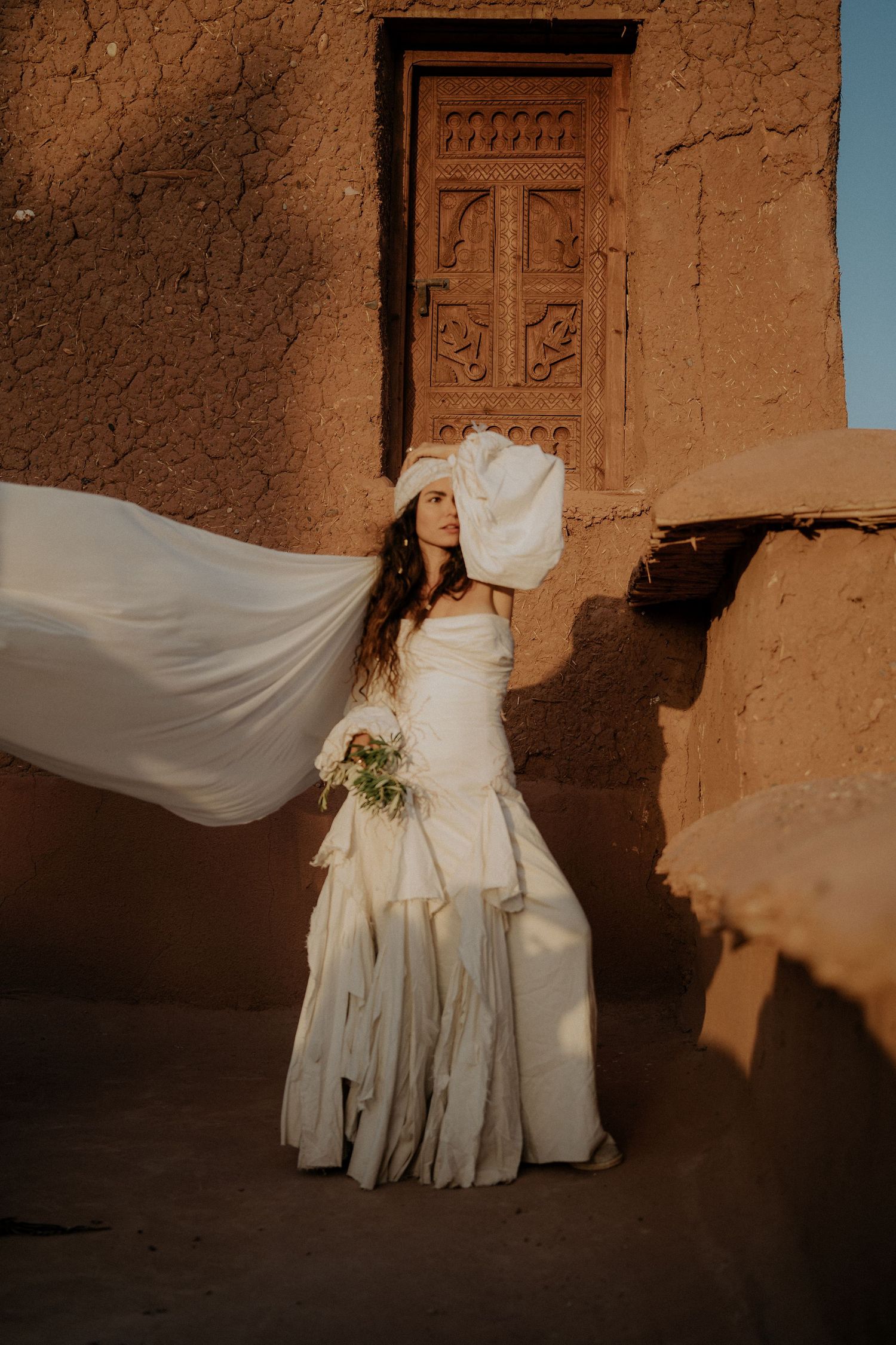 A flowing white wedding dress billows in the wind against an adobe wall with an ornately carved wooden door in Morocco.