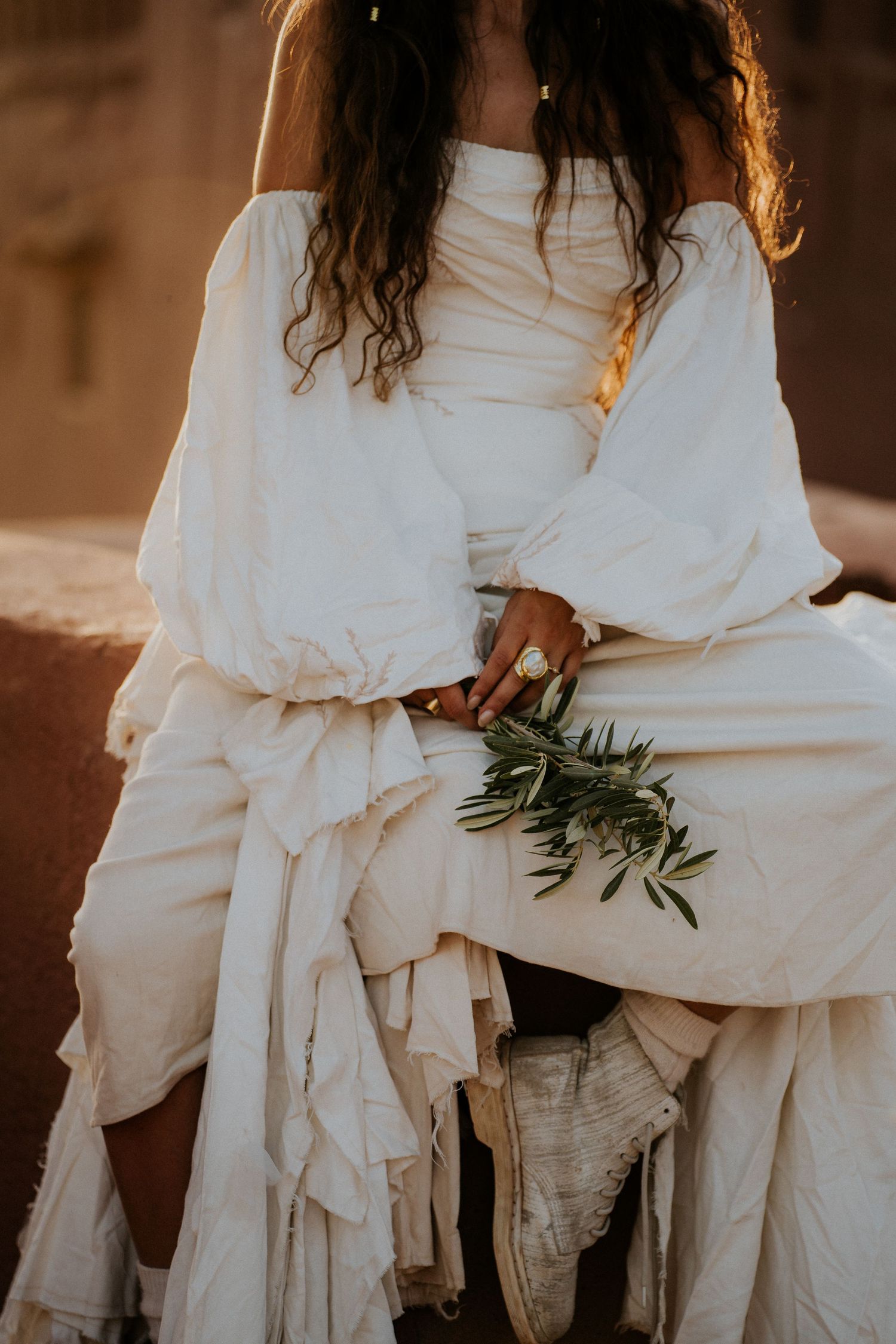 A flowing white dress with olive branches creates an ethereal bridal detail shot in warm sepia tones.