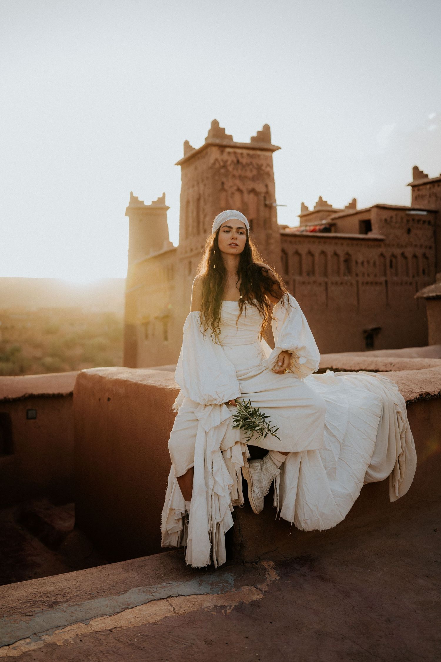 Ethereal desert photoshoot at sunset features flowing white dress against ancient Moroccan kasbah architecture in warm light.