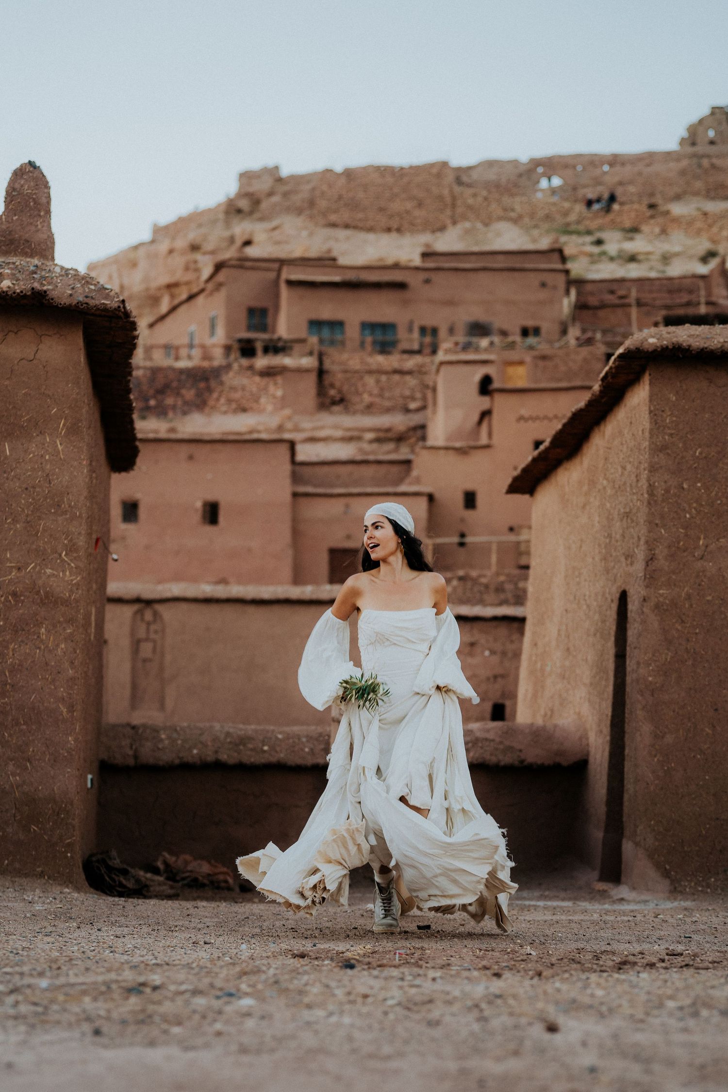 Person in flowing white dress sits near traditional clay buildings in a desert village setting.
