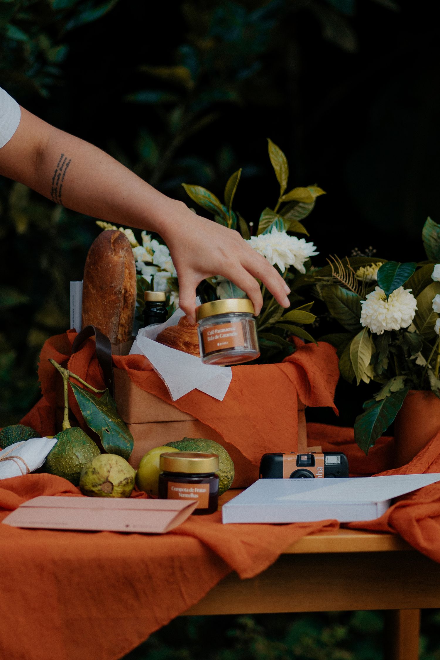 Hand reaching for jar on wooden table with orange cloth and botanical styling.