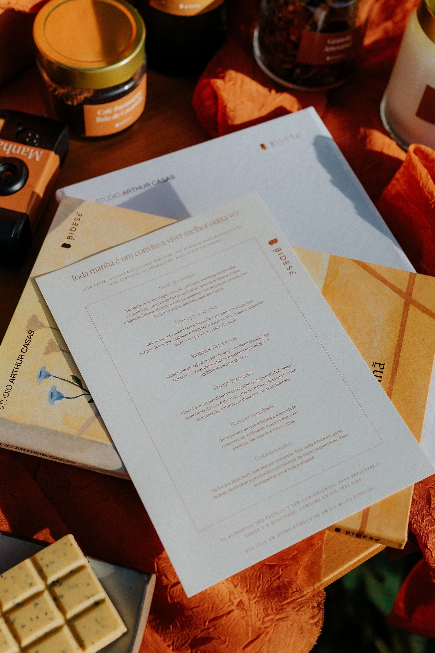 Documents and papers arranged on an orange textile surface with jars and a chocolate bar.