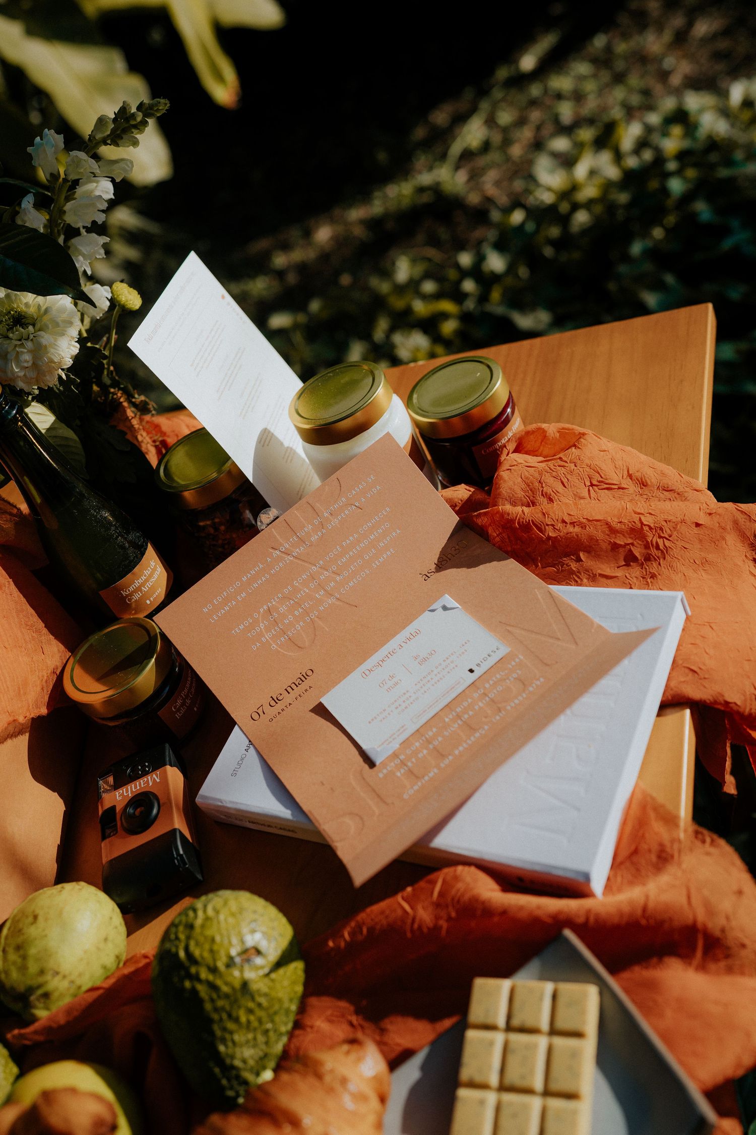 Flatlay of documents, chocolate bars, fruits and jars arranged on an orange textile in natural light.