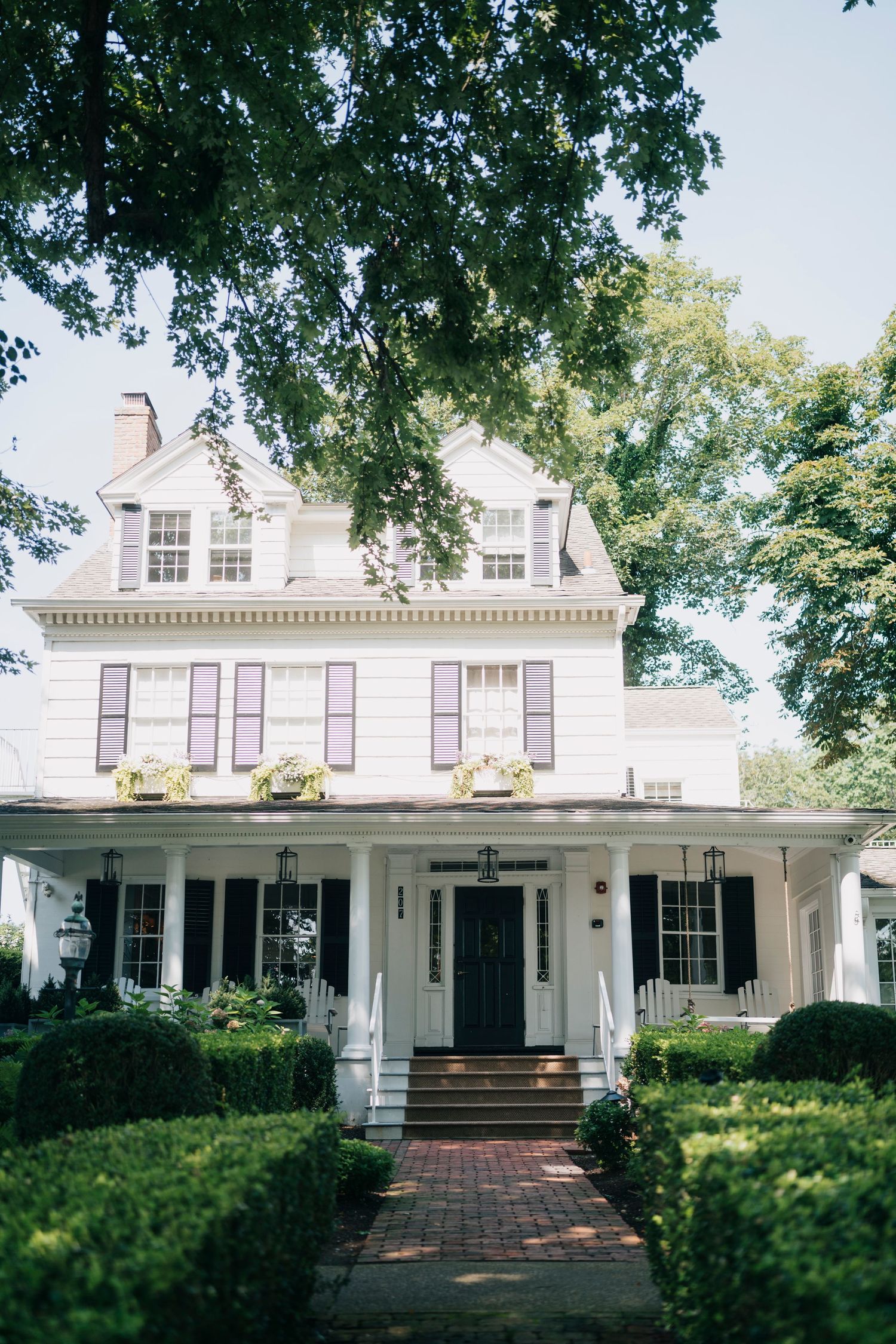Classic white Southern plantation-style house with columned porch and black shutters surrounded by mature trees.