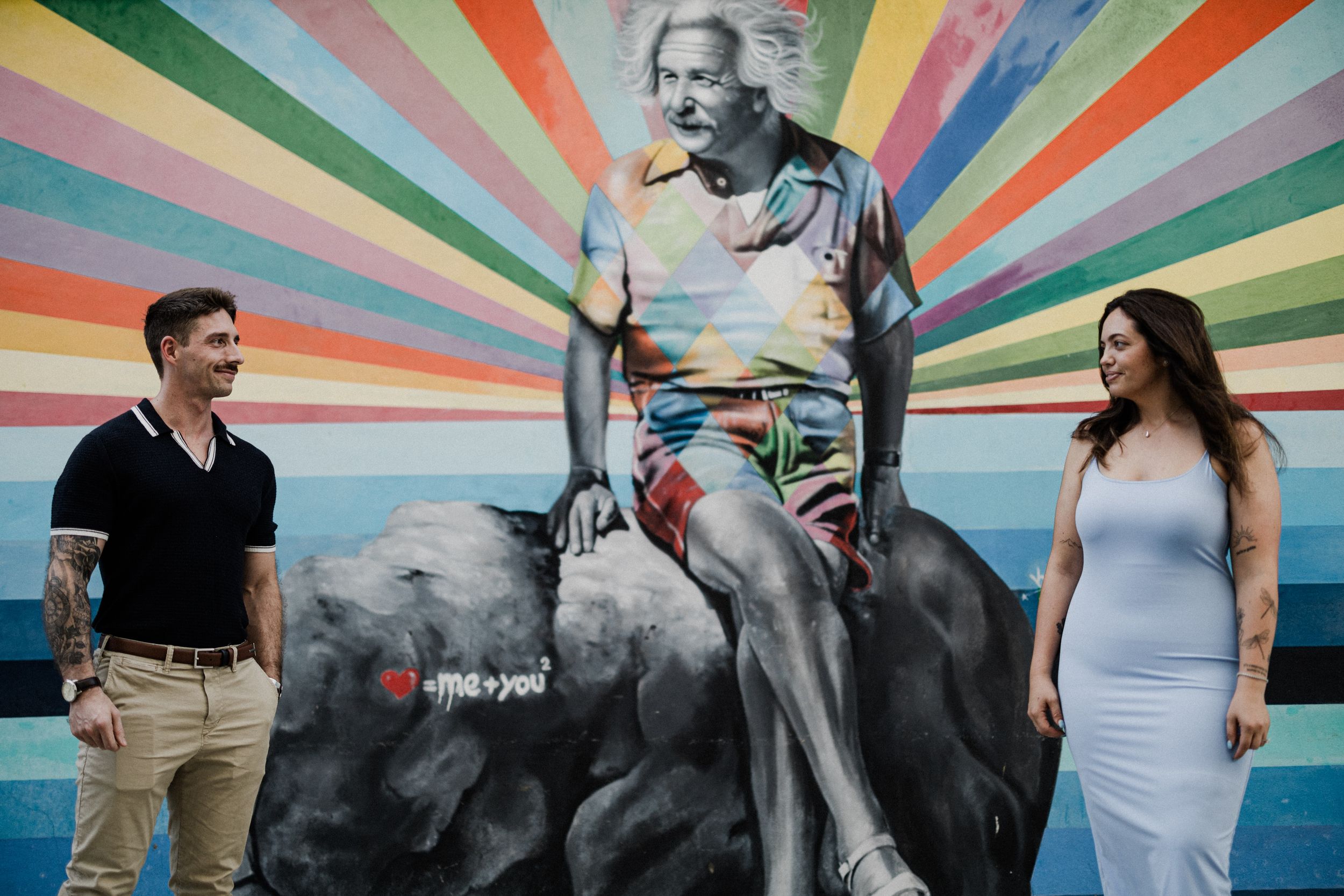 A couple poses in front of a vibrant rainbow starburst wall mural in an urban setting for wedding or engagement photos.