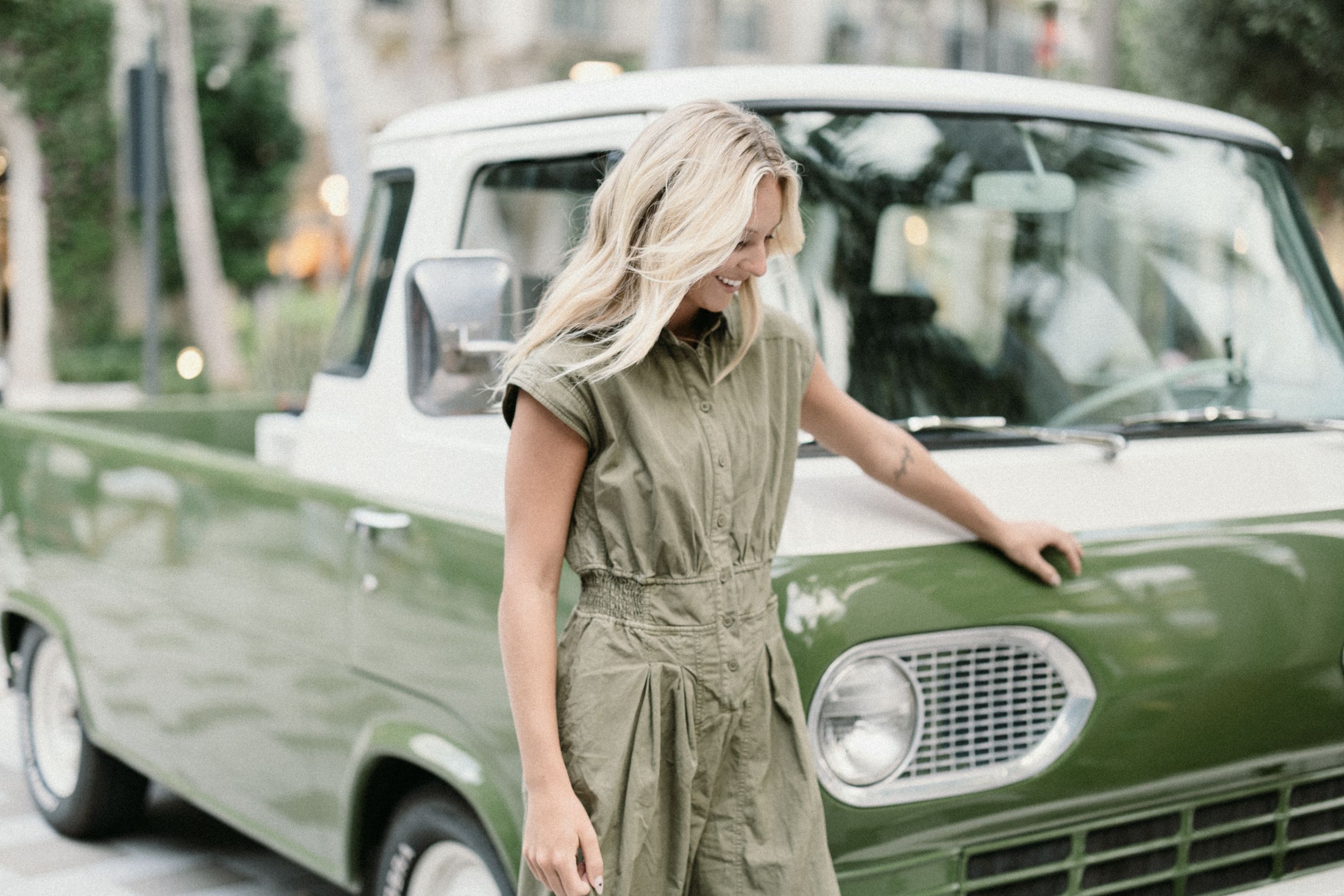 Woman in olive dress posing next to vintage cars during a sunny outdoor photoshoot.
