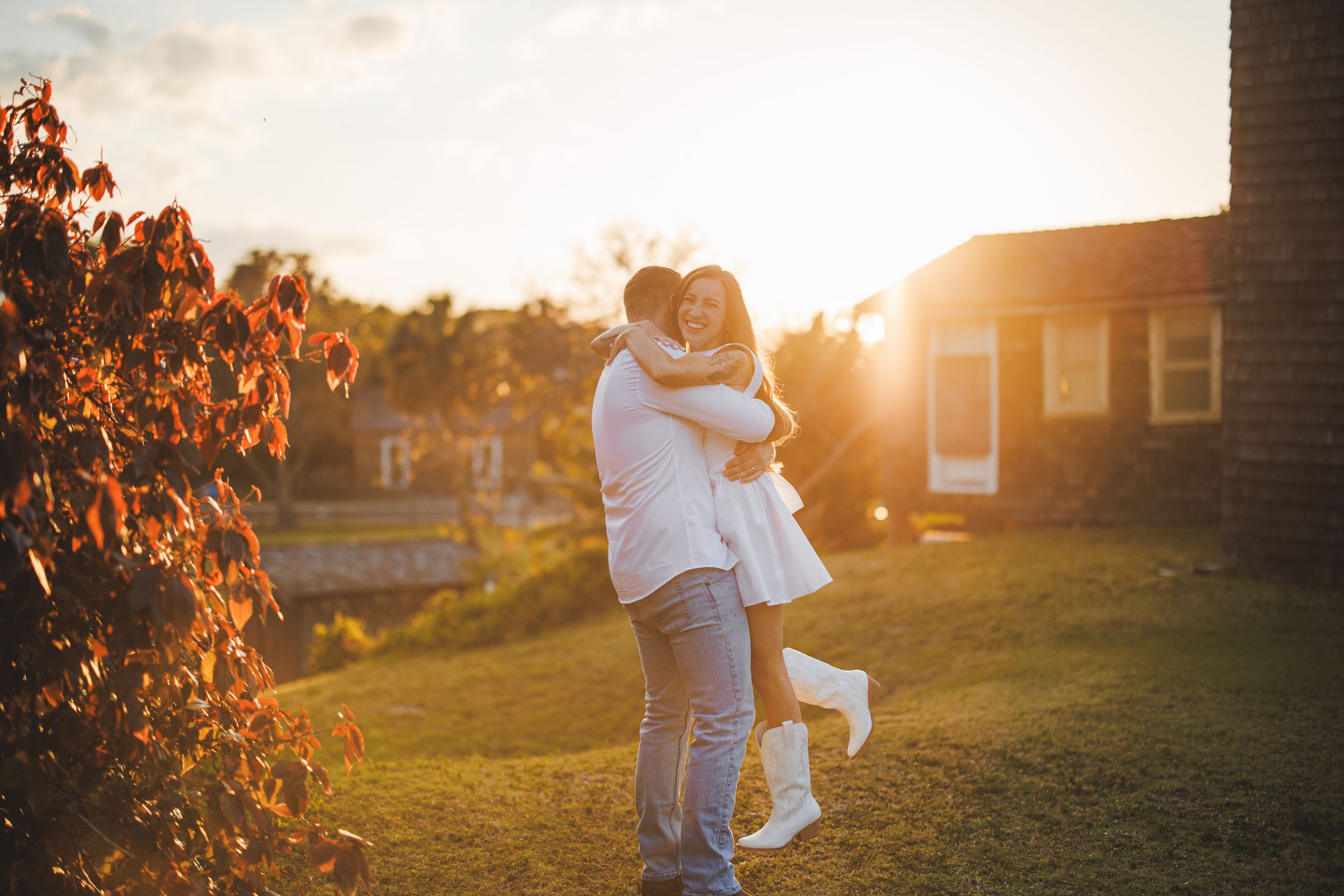 A couple embraces in a backyard at golden hour as warm sunlight filters through trees creating a romantic silhouette.