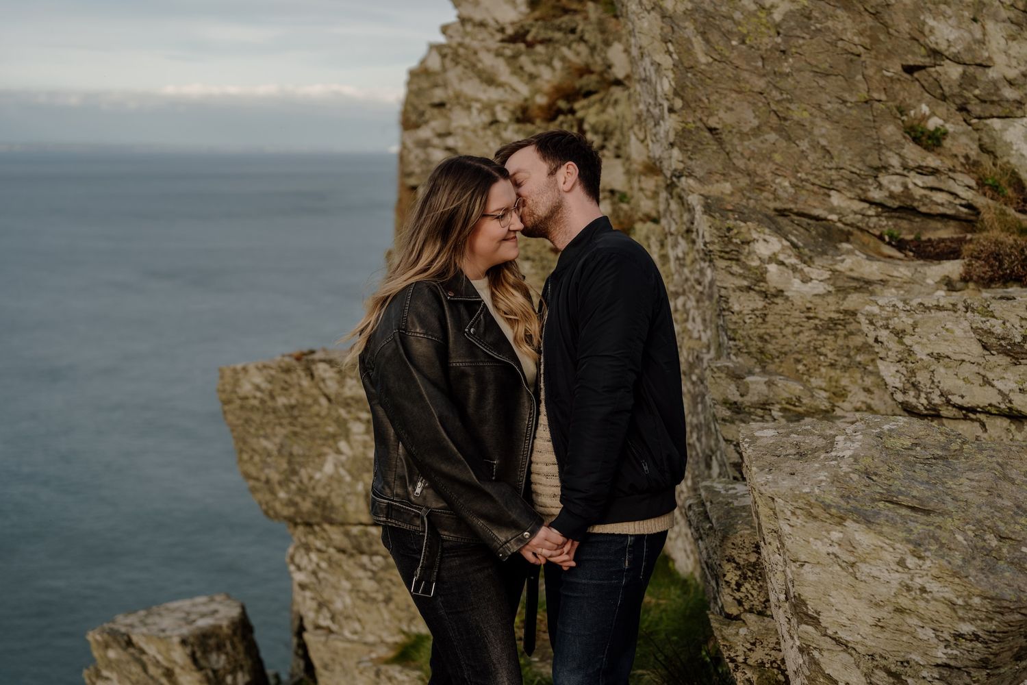 A romantic couple shares an intimate moment on rocky coastal cliffs overlooking a misty ocean.