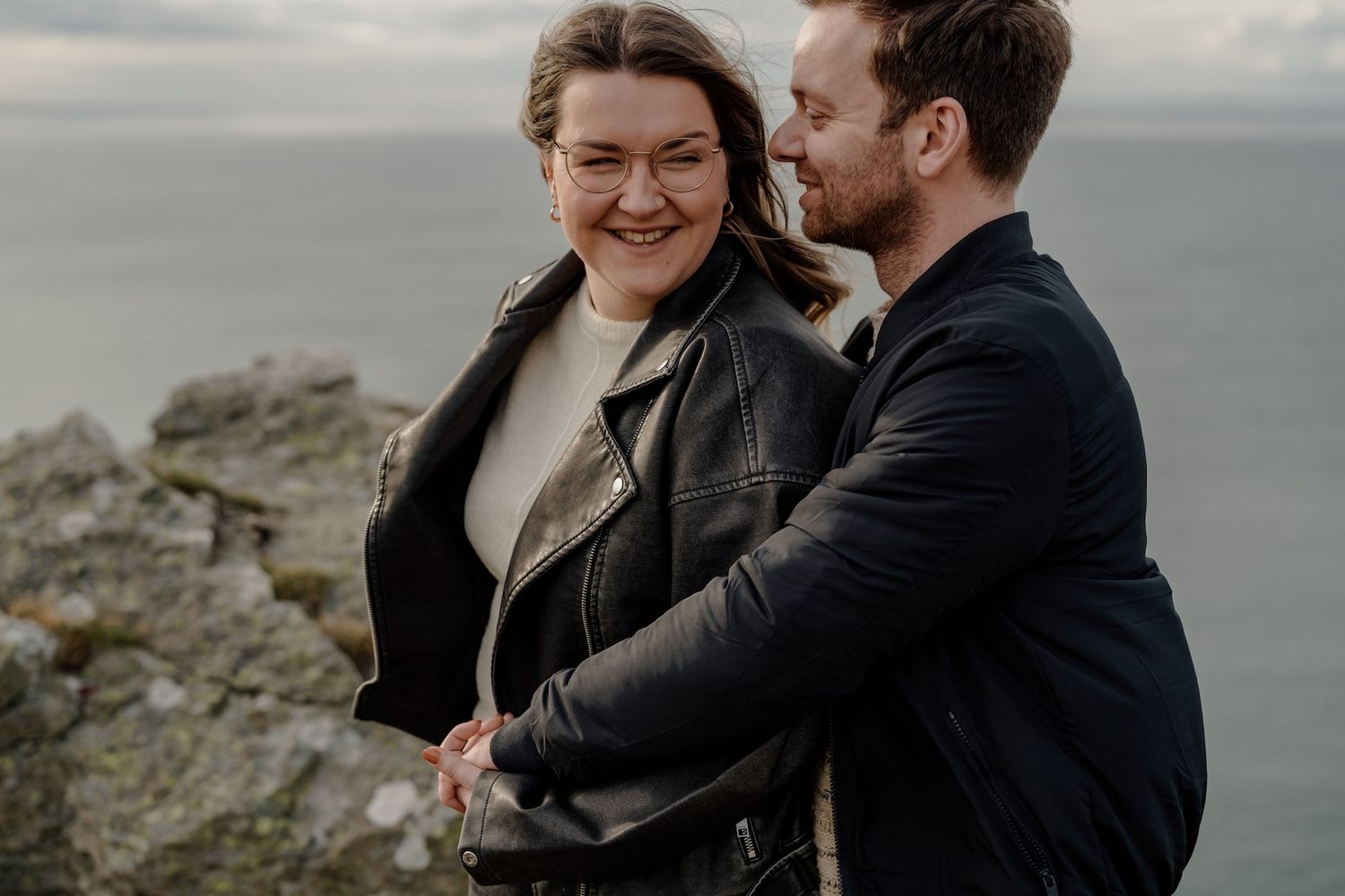 A romantic couple embraces against a moody coastal backdrop with dramatic clouds and rocky cliffs.