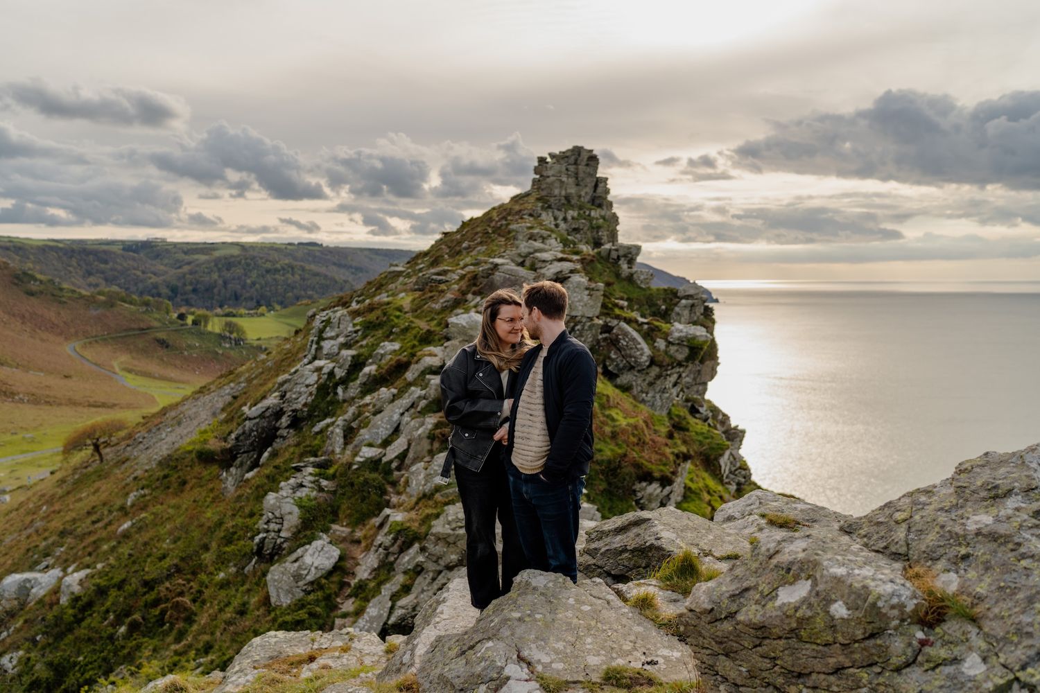 Two people share an intimate moment atop dramatic coastal cliffs overlooking the ocean at sunset.