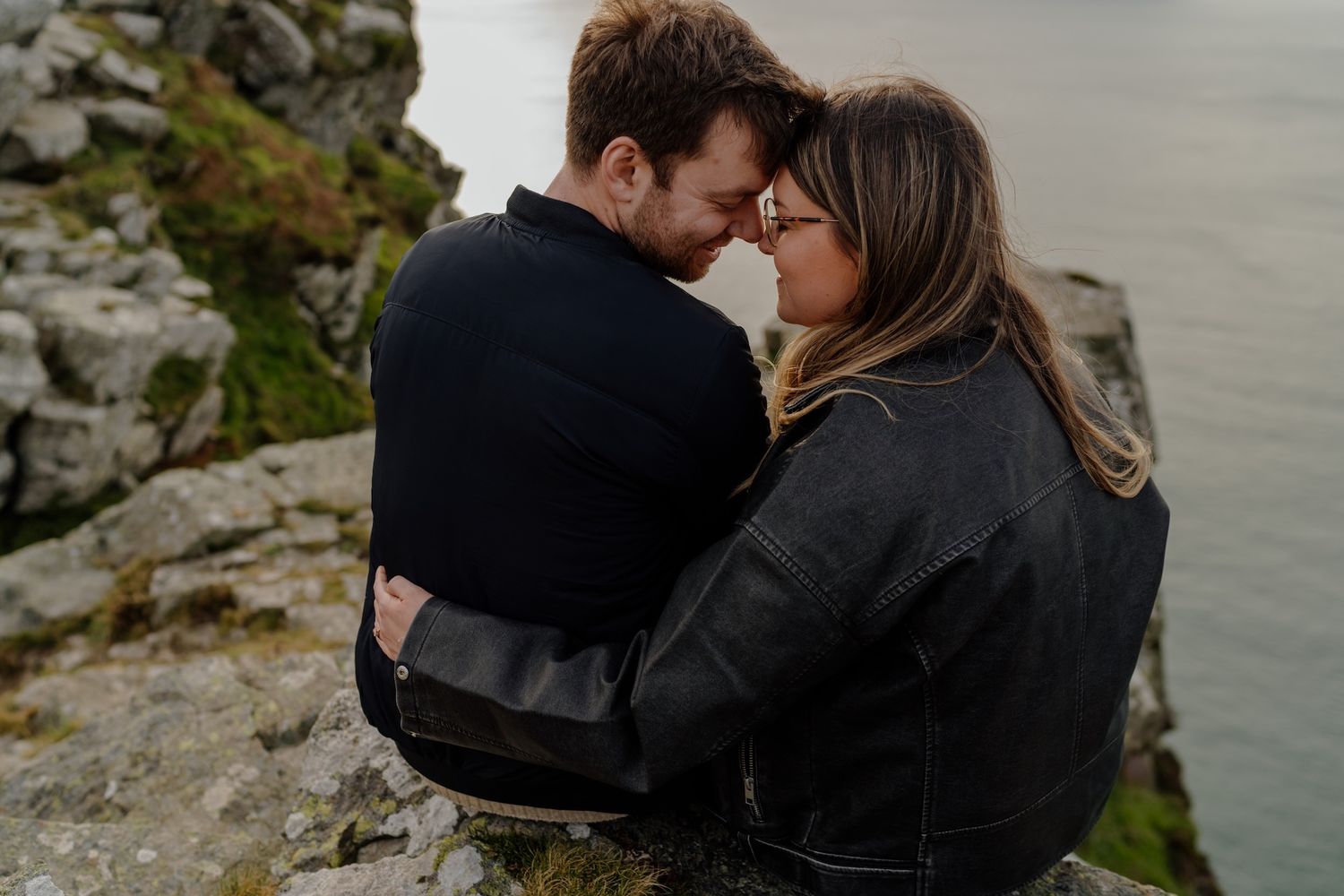 Two people embracing on a cliff edge with dramatic coastal scenery and moody skies in the background.