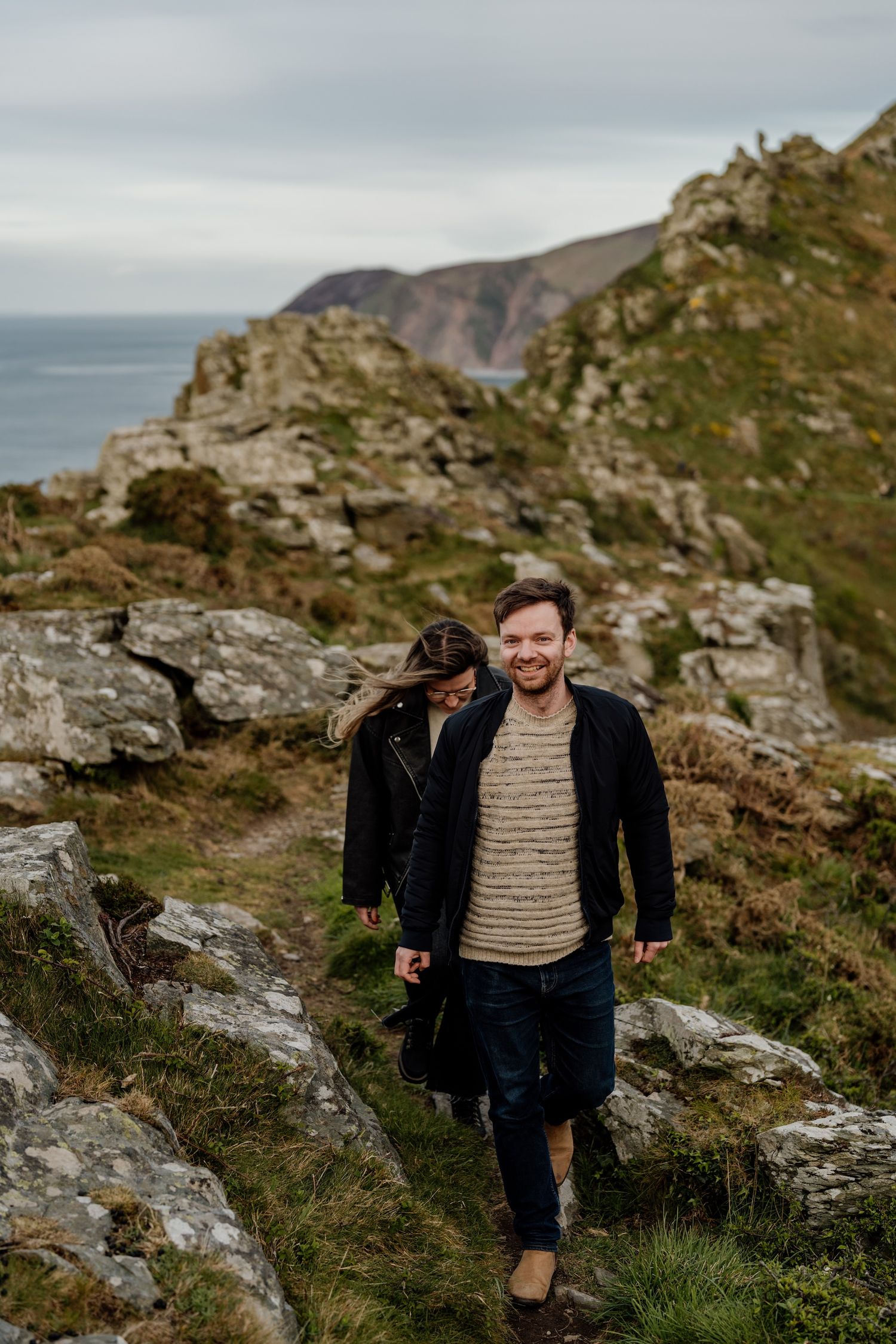 Two people hiking along rugged coastal cliffs with ocean views on an overcast day.