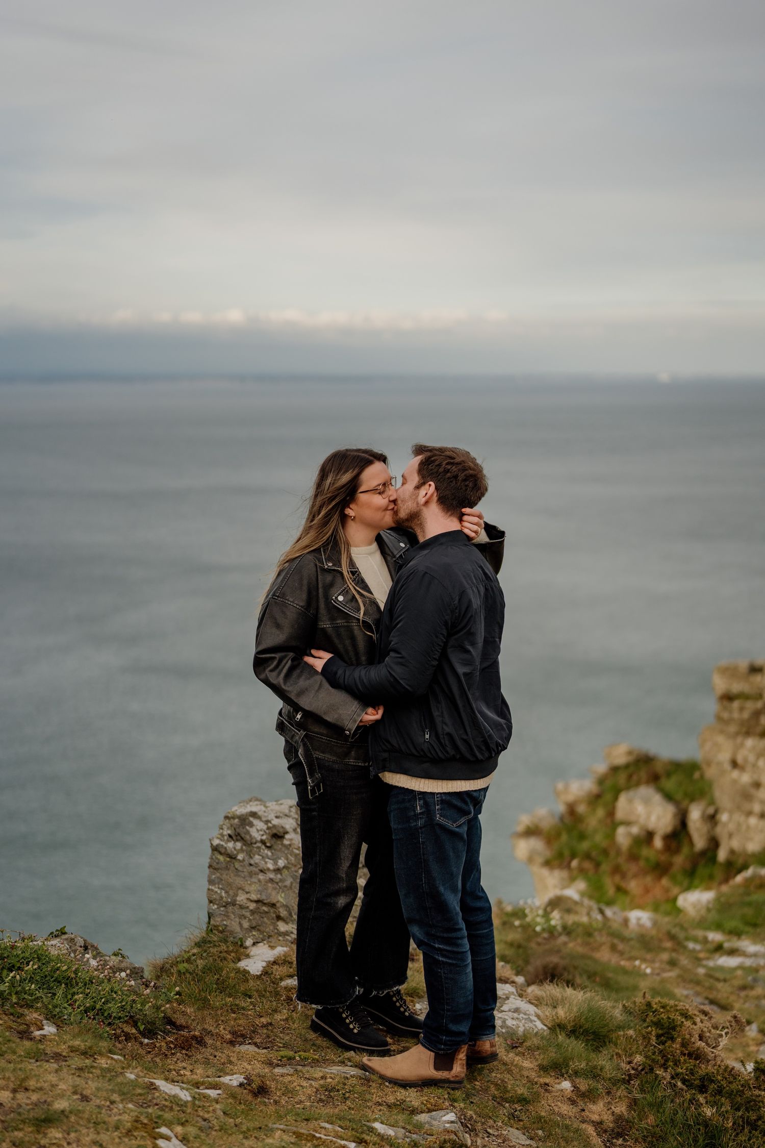 Couple sharing tender moments on windswept coastal cliffs overlooking a misty ocean.