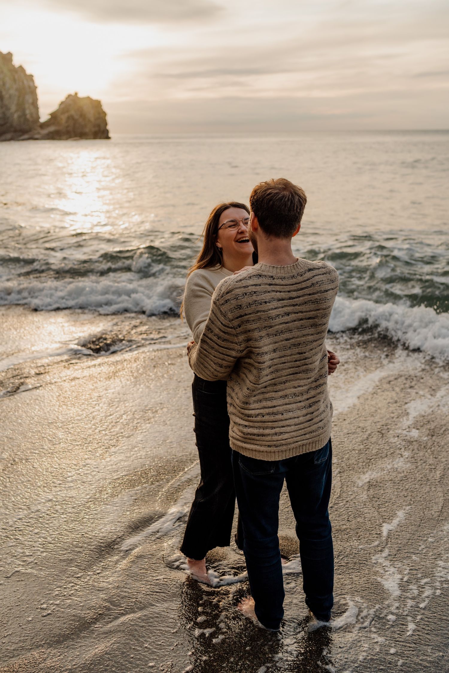 A couple embraces at sunset on a beach with waves crashing around their feet.