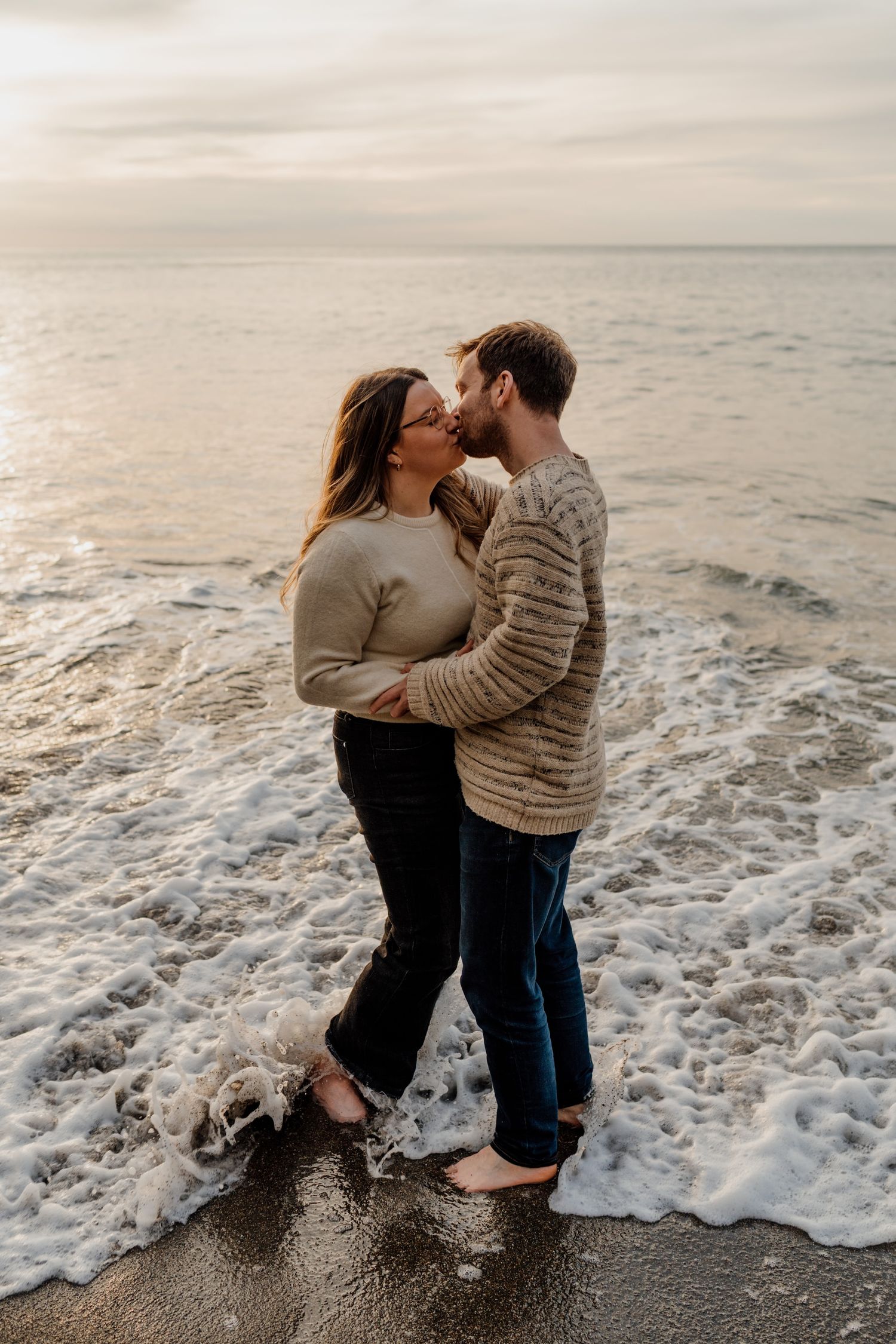 Couple embracing on a beach as ocean waves crash behind them during golden hour.