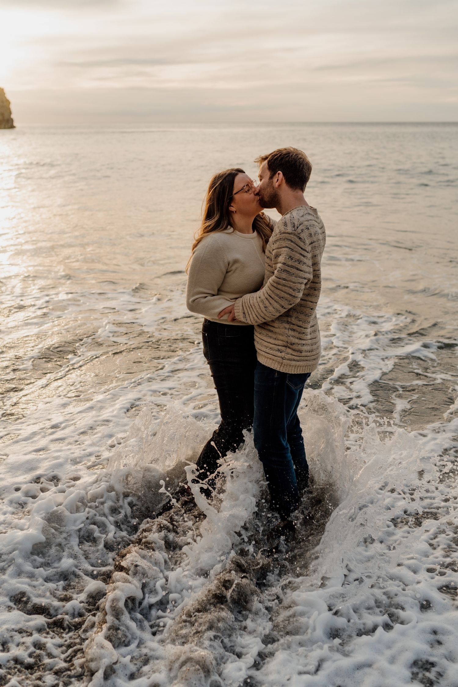 Two people embrace in the ocean waves as water splashes around them during a romantic sunset photoshoot.