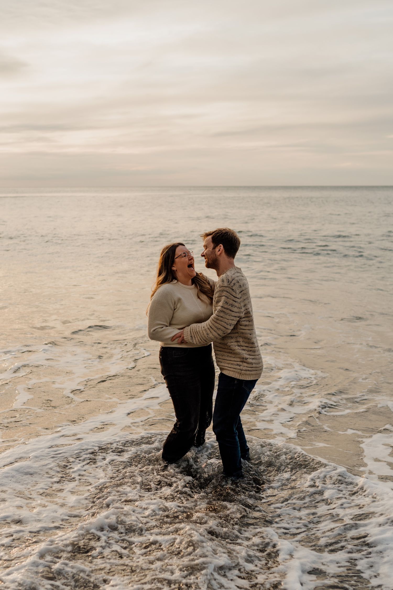 Couple playing and laughing together while standing in shallow ocean waves at sunset.