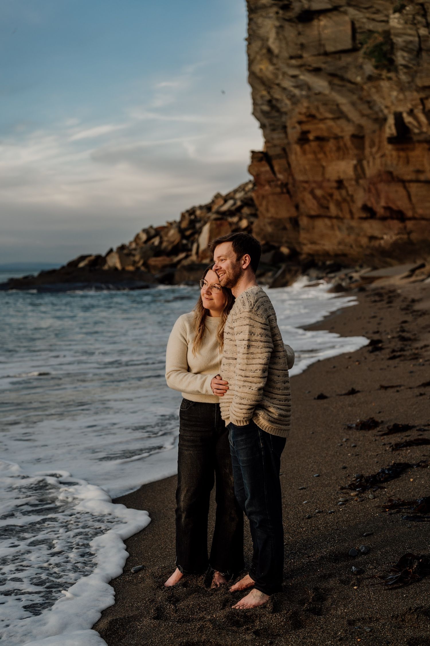 Couple sits together on dark rocky beach as waves crash along the shoreline at dusk.