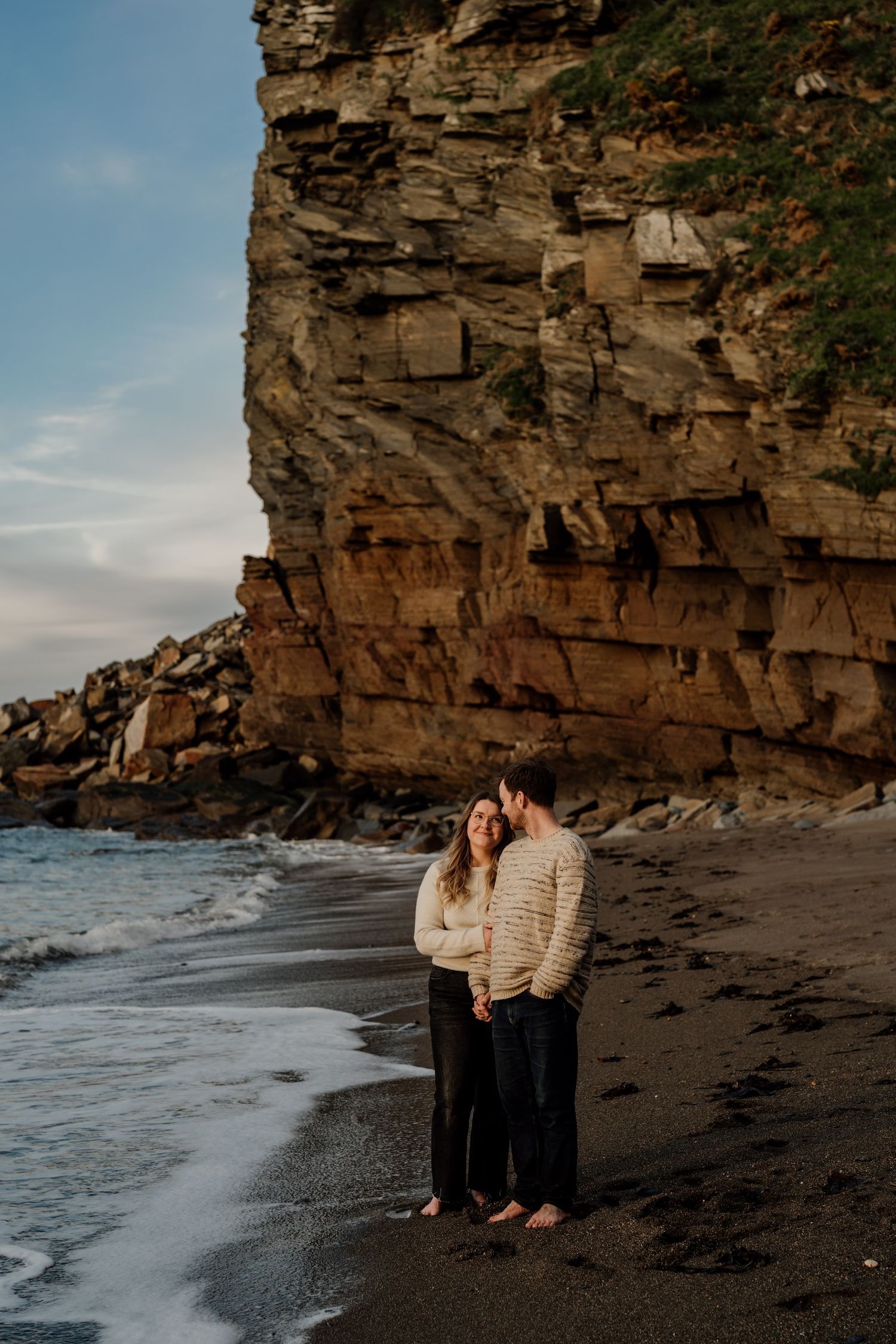 Couple sharing romantic moments on a beach with dramatic rocky cliffs in the background.