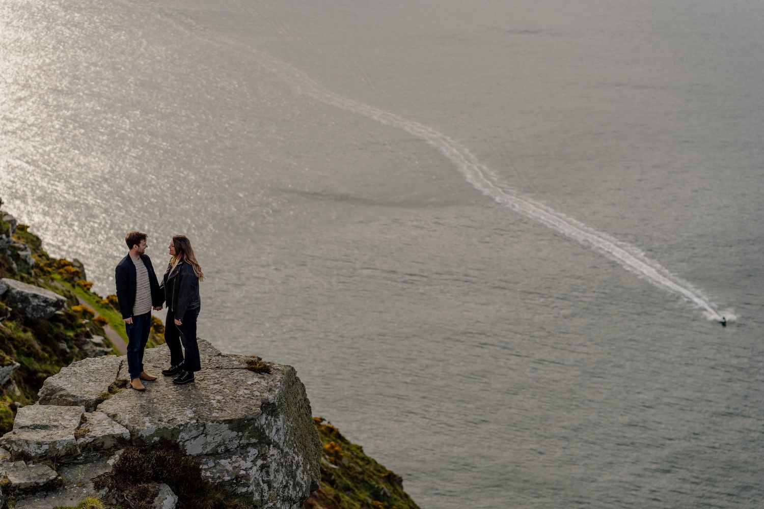 Two figures stand on a dramatic rocky cliff overlooking a misty ocean with a boat leaving a wake trail below.