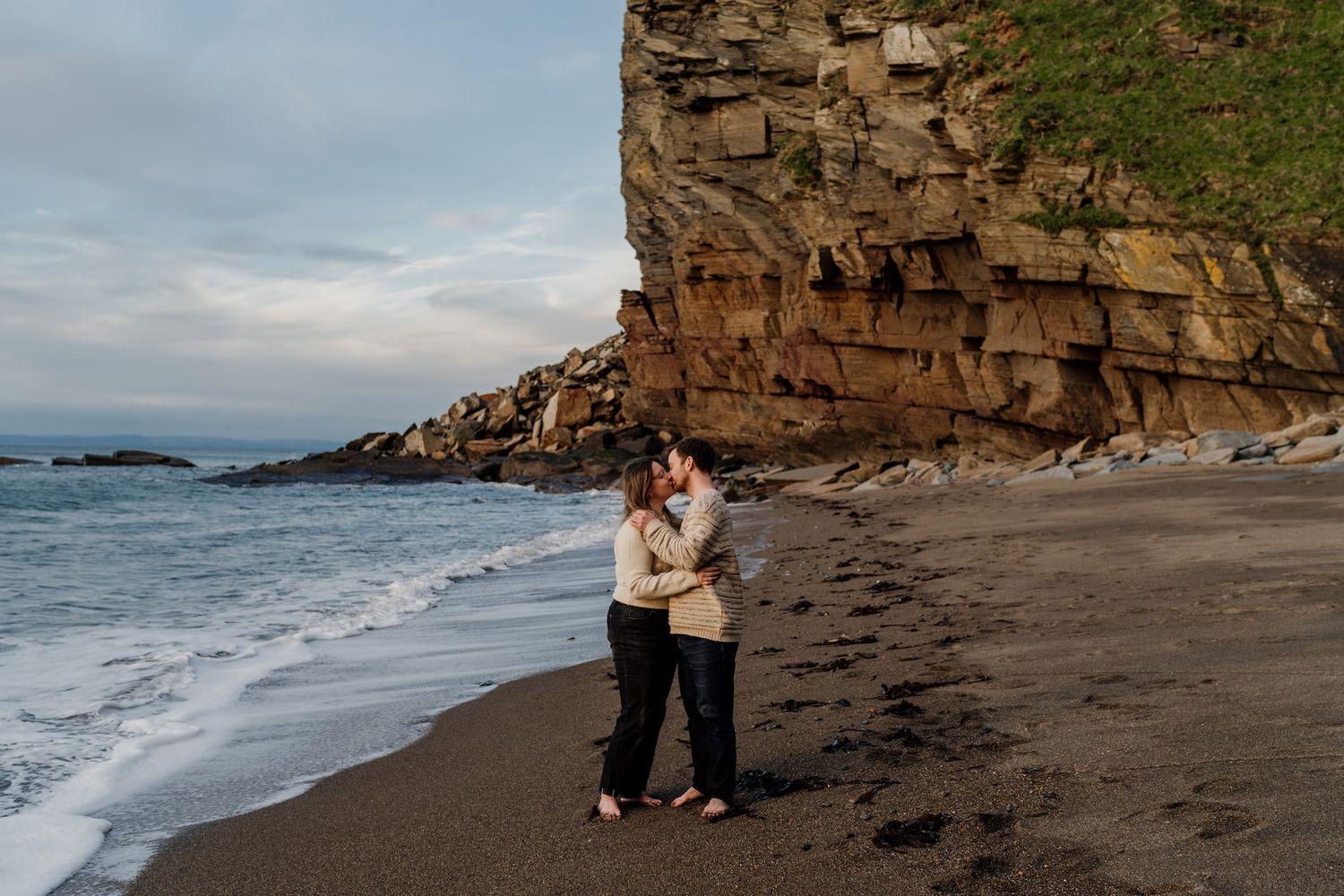 A couple embraces on a secluded beach with dramatic cliffs and ocean waves in the background.