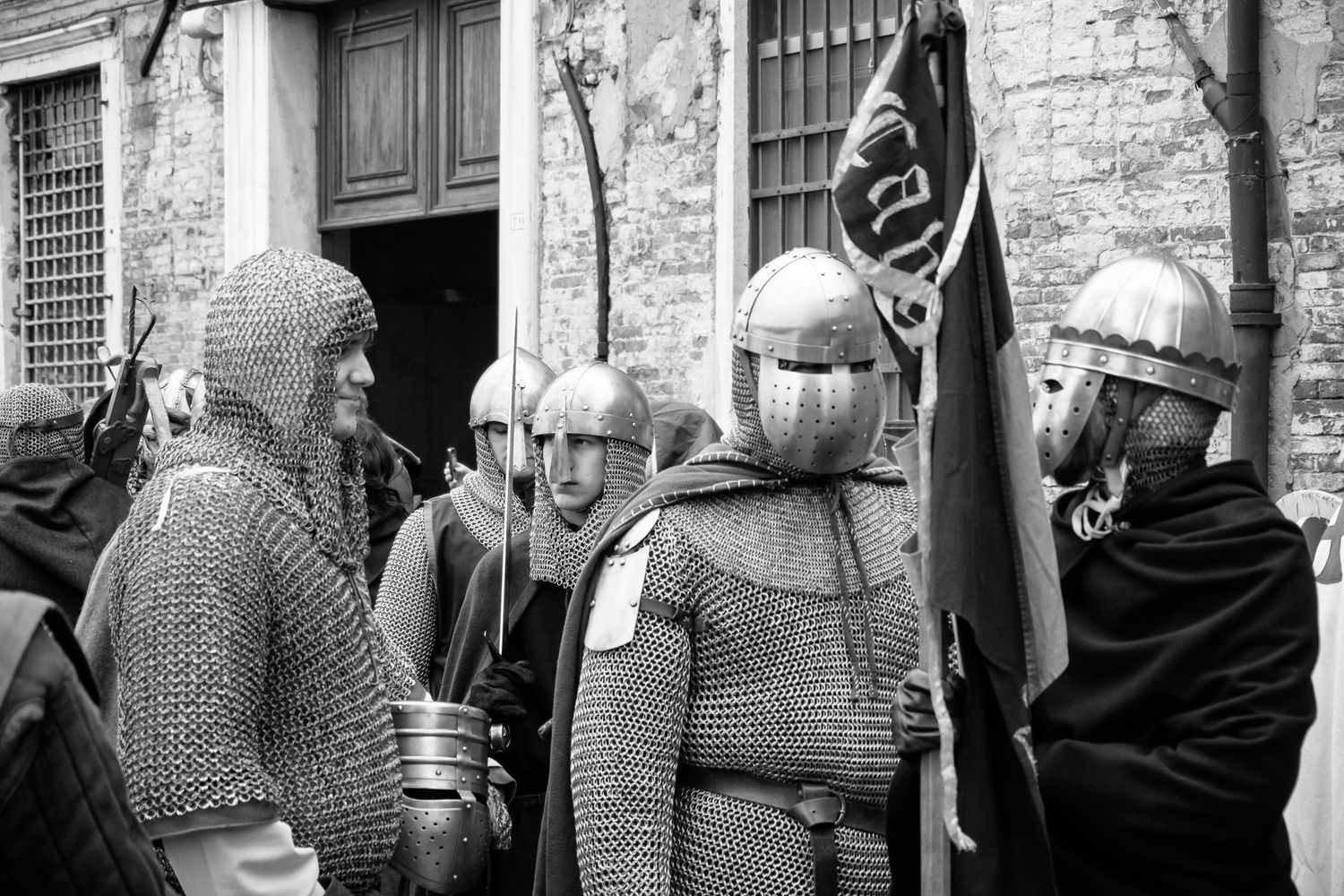 Medieval reenactors in chainmail armor and helmets gather around a black flag at a historic building entrance.