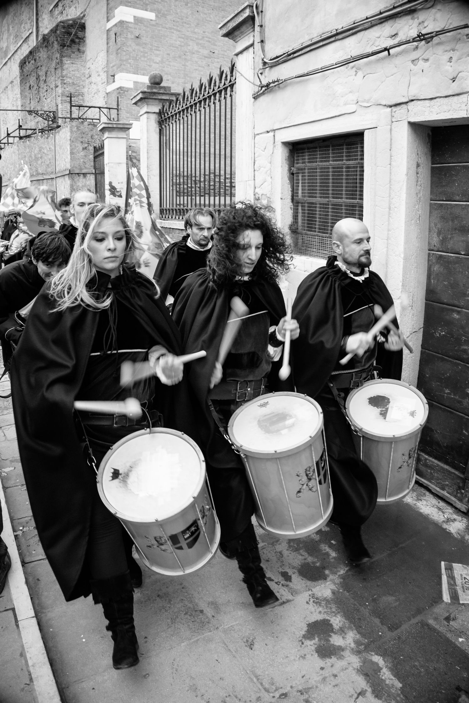 Drummers in black capes perform during a street festival procession in black and white photography.