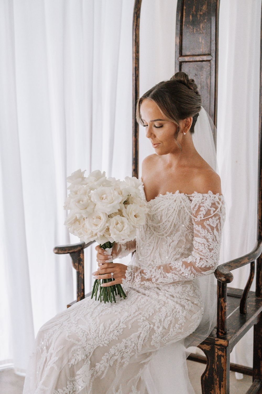 Elegant bride in detailed lace wedding gown with intricate details sits gracefully in antique wooden rocking chair.