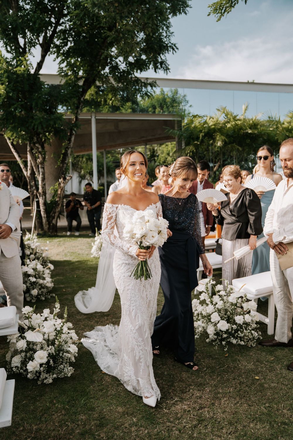 Elegant outdoor wedding ceremony with white floral arrangements and guests gathered on lawn under trees.