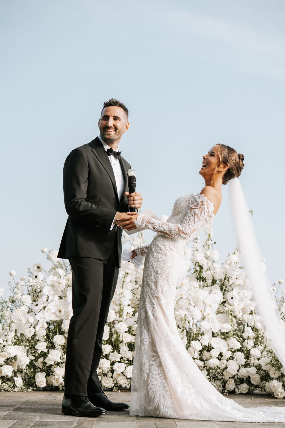 Newlyweds laughing and embracing in front of a beautiful white floral wall backdrop at an elegant wedding ceremony.