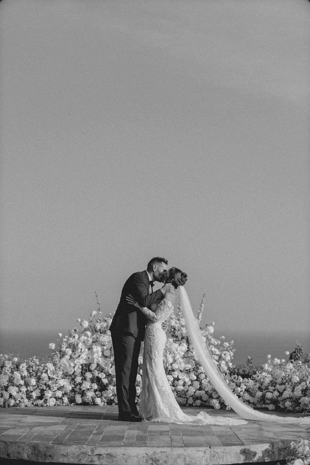 Black and white romantic wedding photo series showing couple in formal attire with floral arrangements against ocean backdrop.