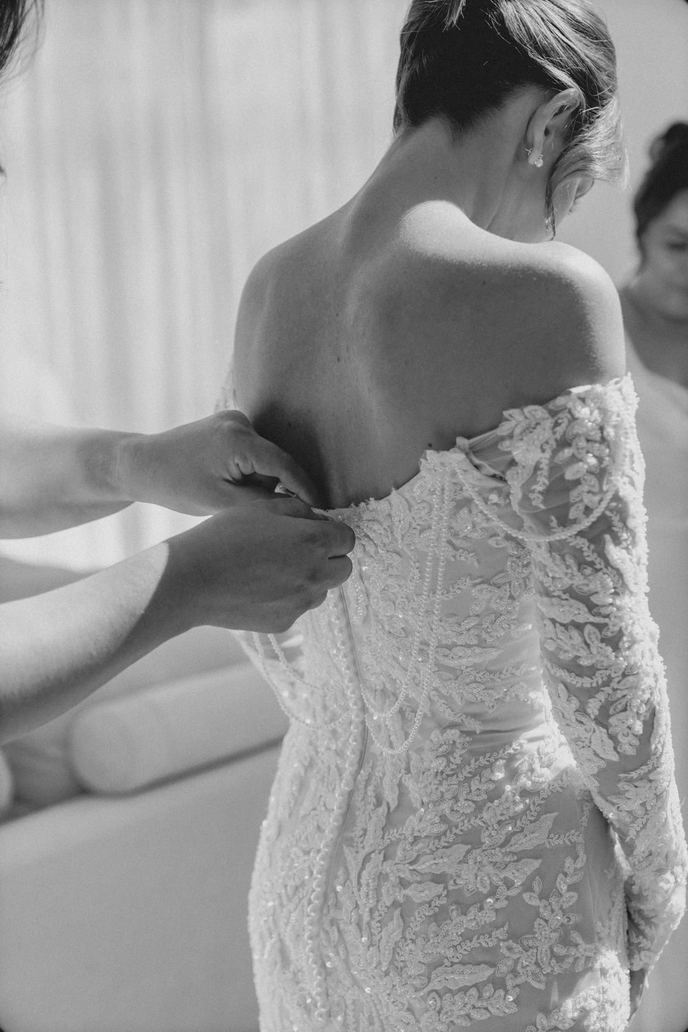 Black and white photo of a bride being fitted into an elegant lace wedding dress with long sleeves.