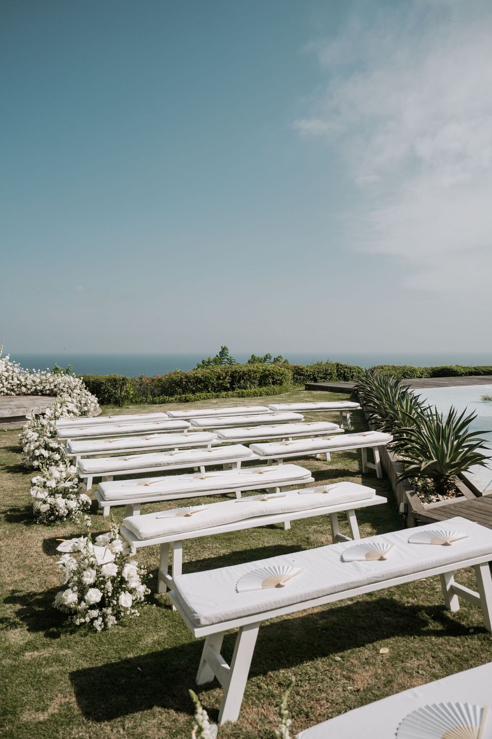 White wooden benches arranged in rows on a grassy cliff overlooking the ocean for an outdoor coastal wedding ceremony.