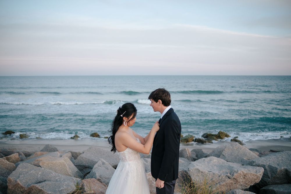A couple in wedding attire stands on a rocky ocean shore with waves crashing behind them during sunset.