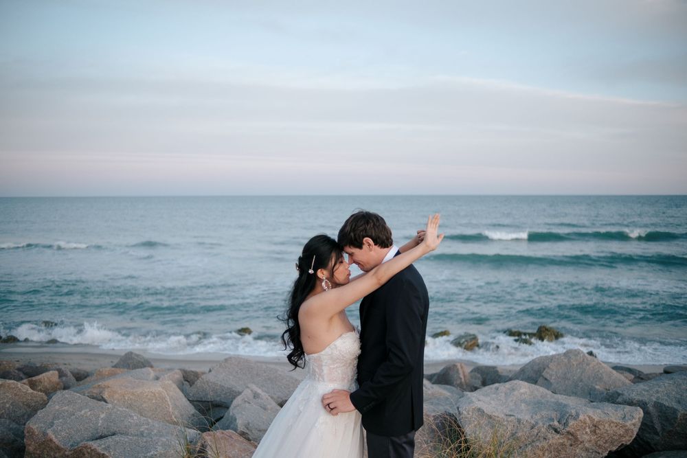 A couple shares an intimate moment against a rocky beach background as waves crash on the shore at sunset.