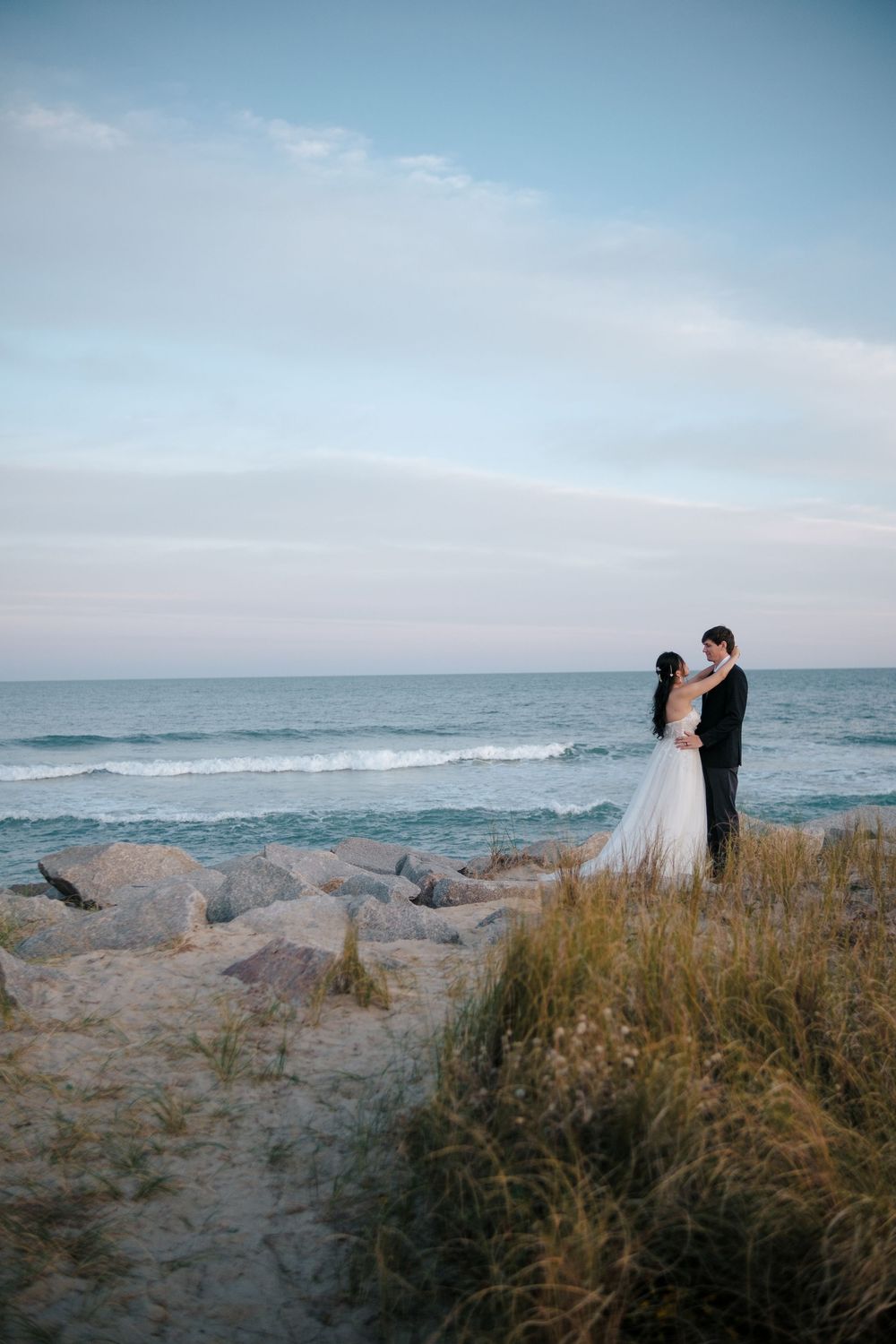 A romantic wedding couple stands on a rocky beach at sunset with ocean waves and beach grass in the foreground.
