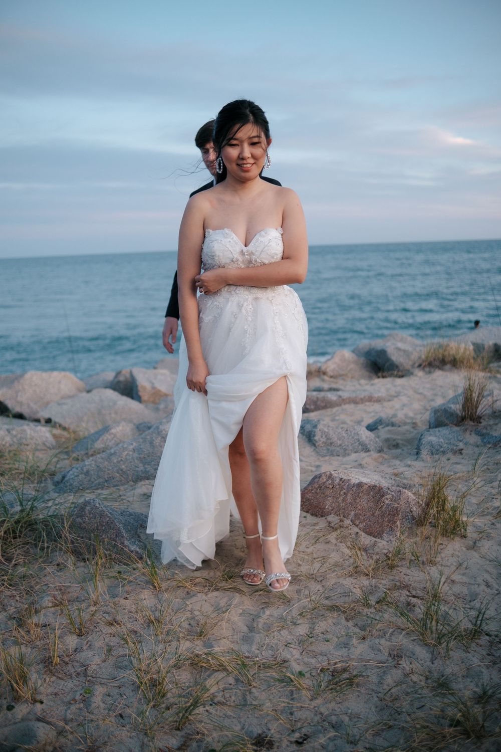 A seaside wedding photo series shows someone in a white gown sitting gracefully among coastal rocks at sunset.