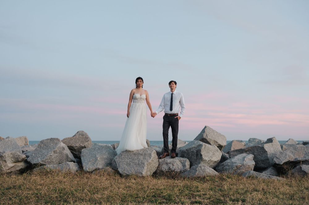 A couple in wedding attire walks along a rocky coastal shoreline during sunset creating a romantic silhouette.