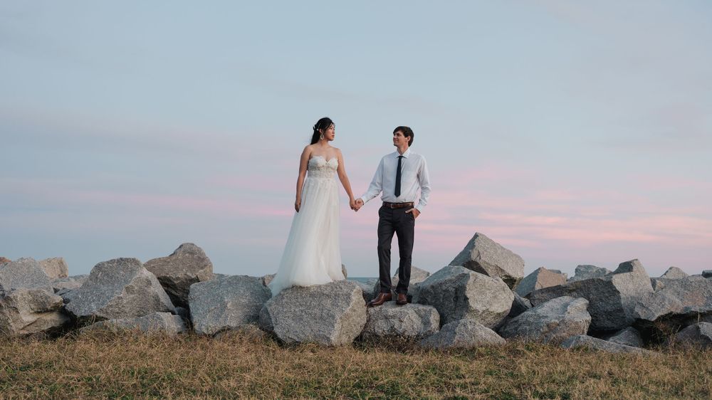 A couple stands on coastal rocks at sunset with mountains in the background.