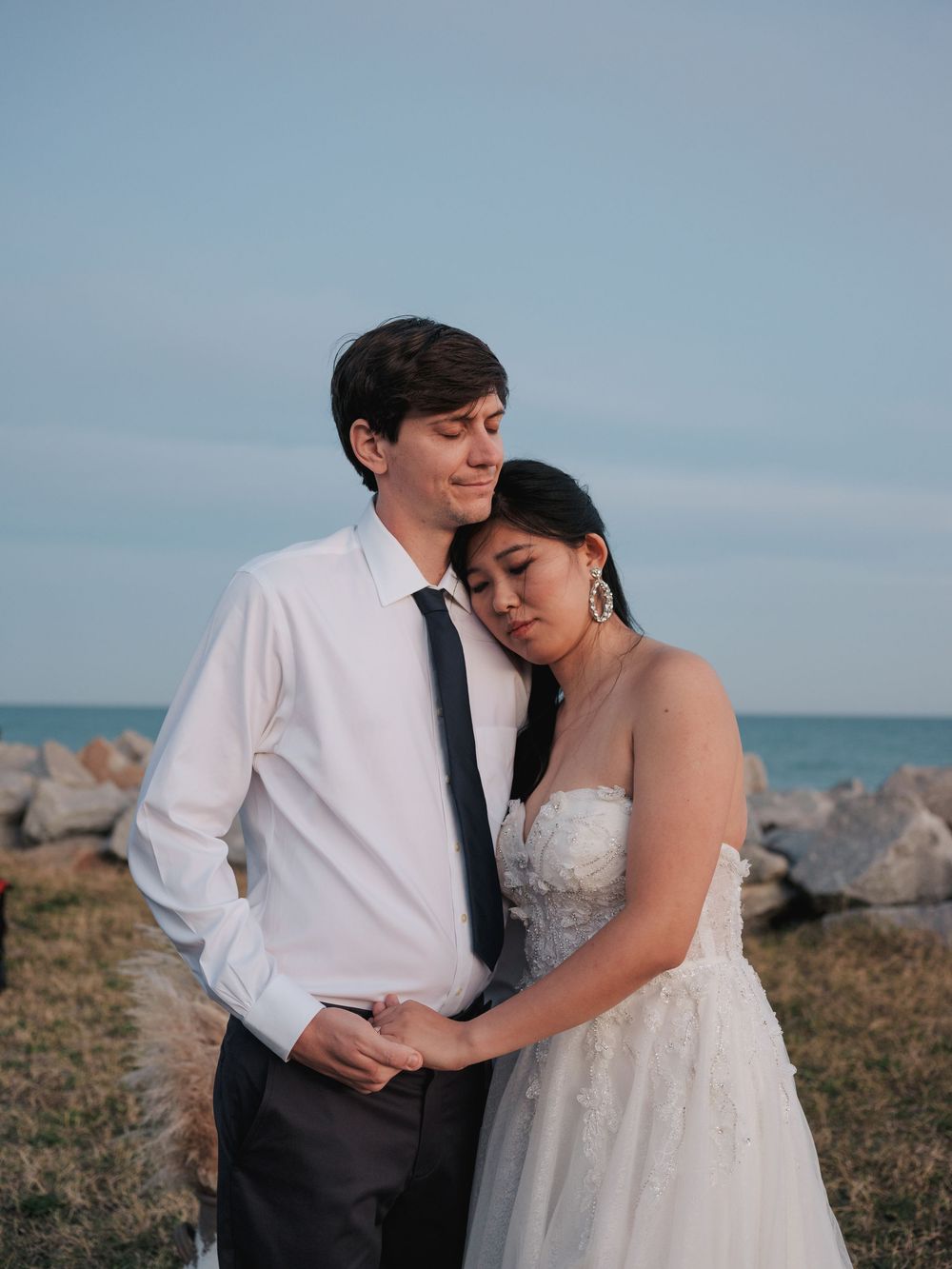 Couple in wedding attire embraces and shares intimate moments by the ocean on a rocky beach.