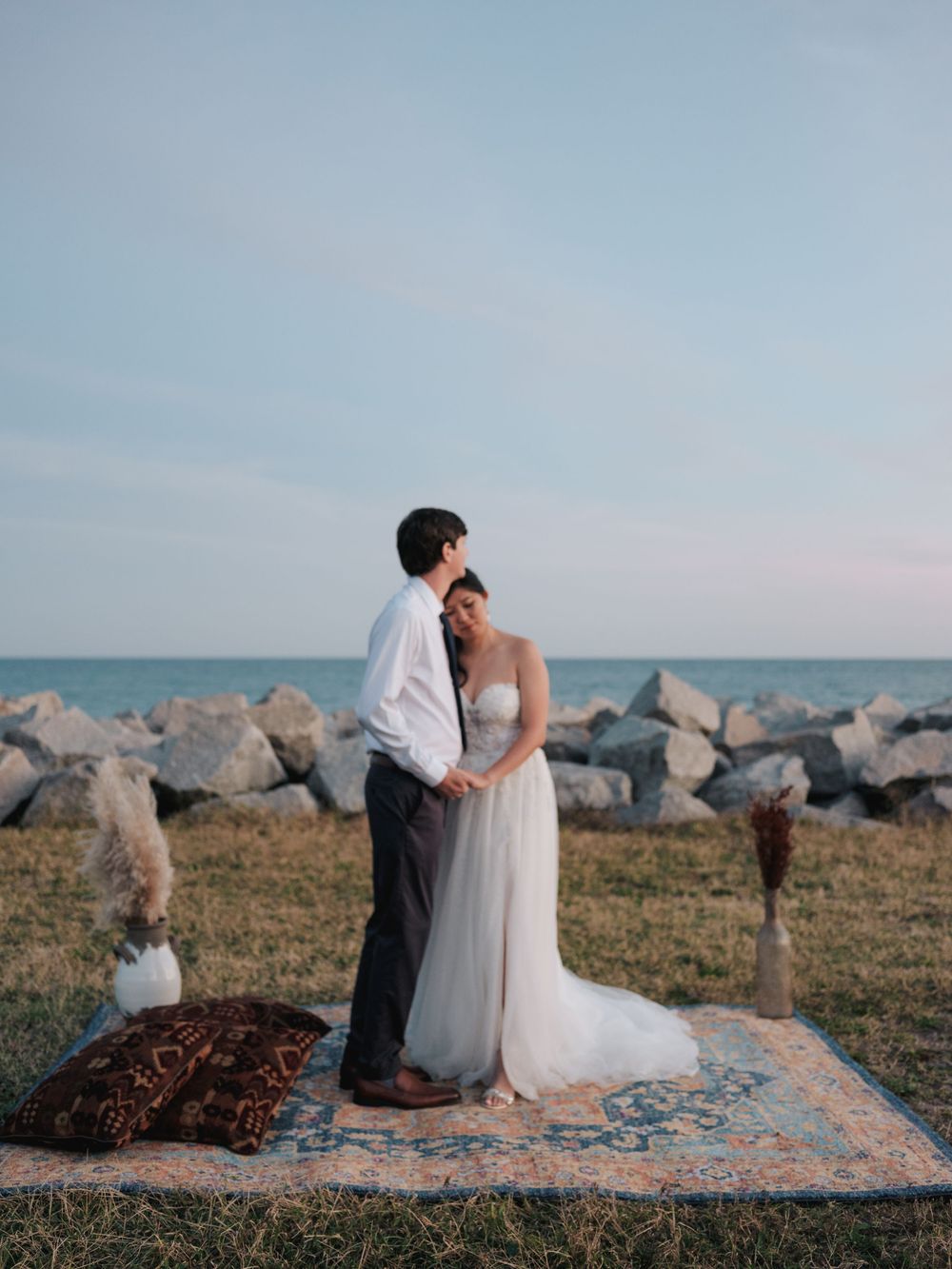 A couple in wedding attire sits beside the ocean on rocky shores during sunset.
