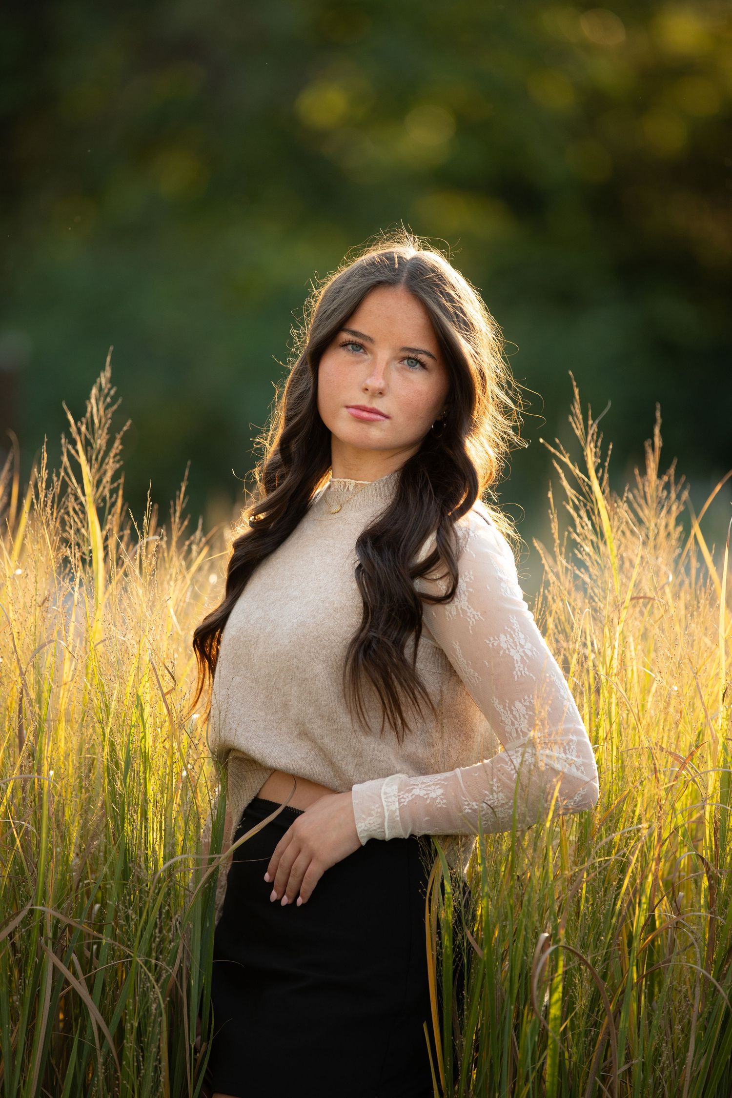 A woman in a cream sweater poses in a sunlit field of golden grass during golden hour.