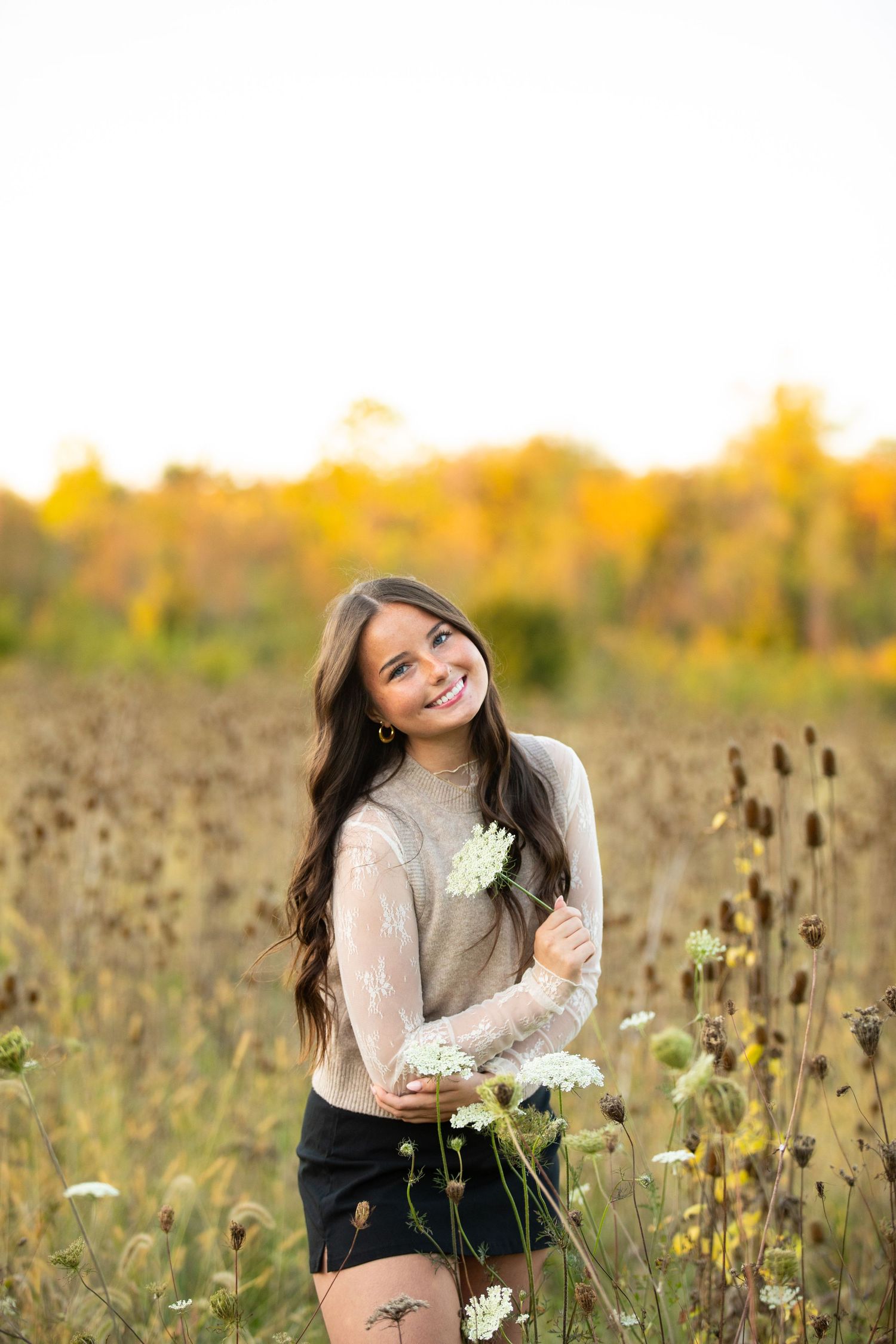 A person in a light sweater poses in a golden autumn meadow with wildflowers at sunset.