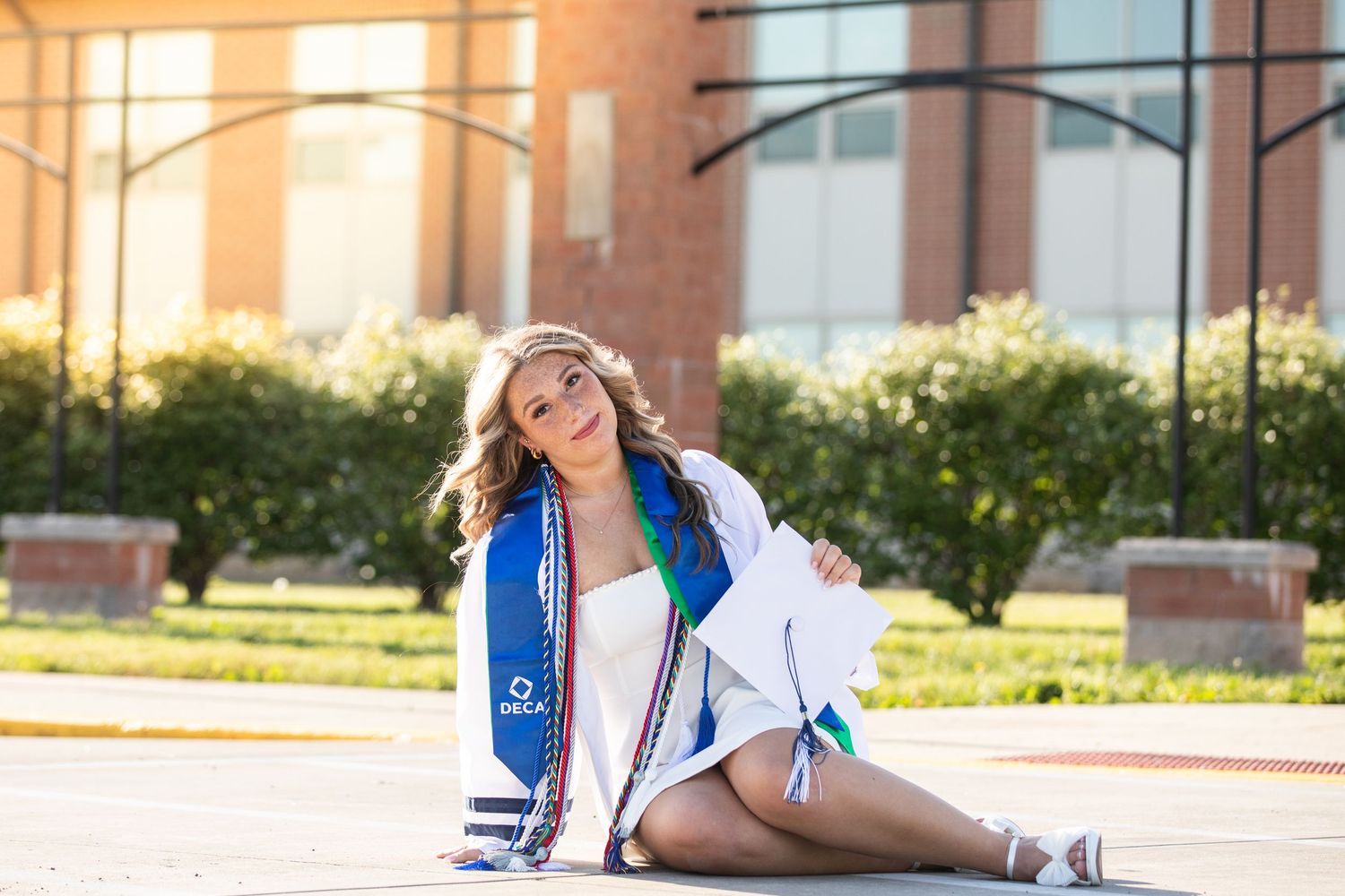 Smiling graduate poses outdoors in white gown with blue sash, sitting on pavement near a brick building.
