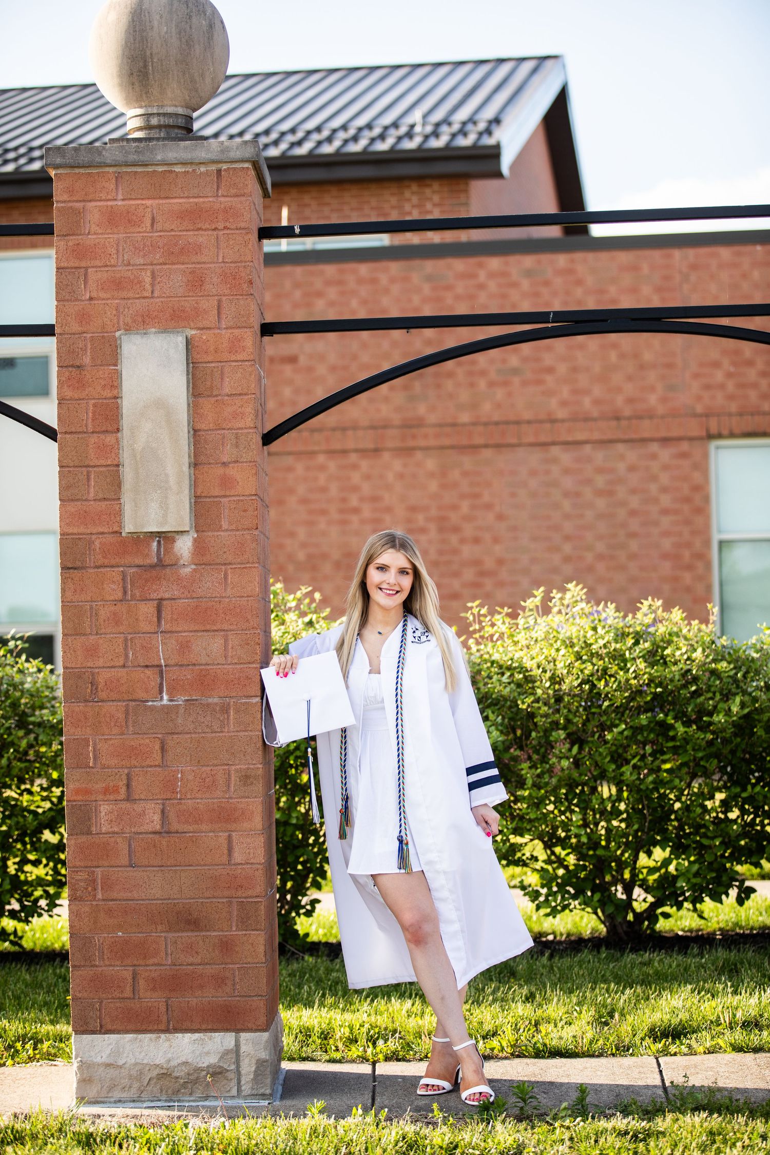 Young woman in white graduation gown holding cap poses by brick pillar on sunny campus grounds.