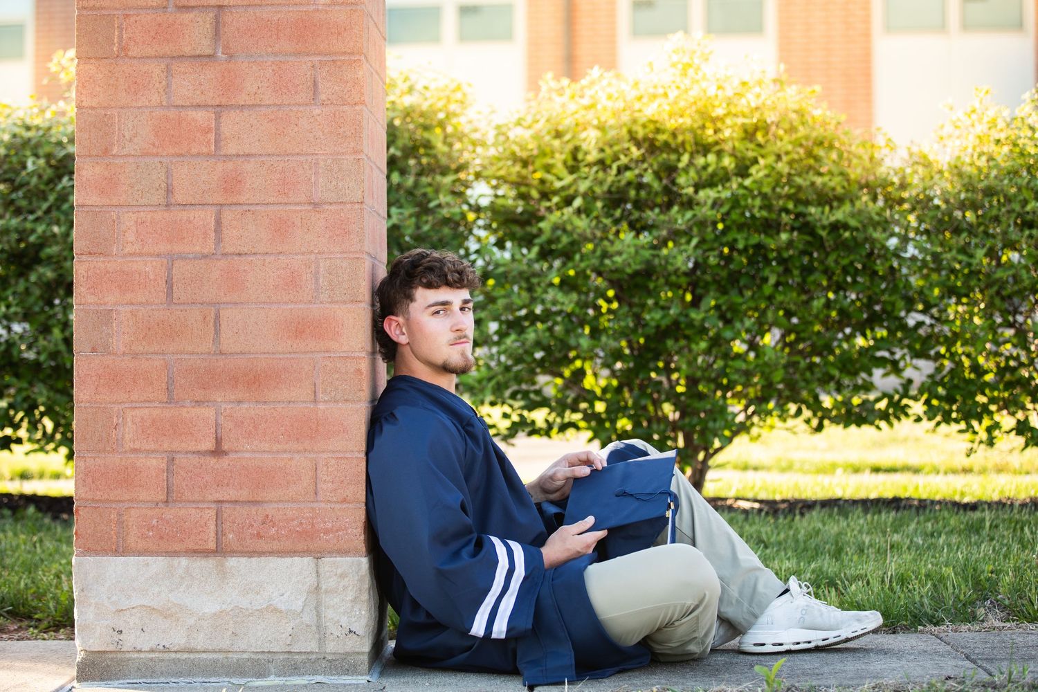 Young male student sitting against brick pillar outdoors, reading a tablet on a sunny campus day.