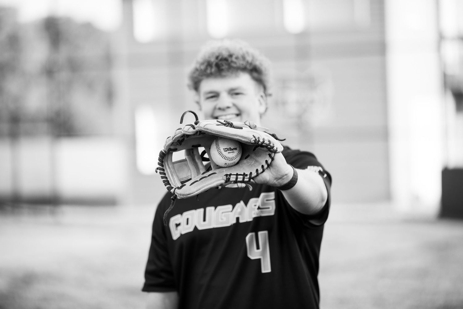 Young baseball player in black Courage jersey holding glove up to mouth, smiling, black and white photo.