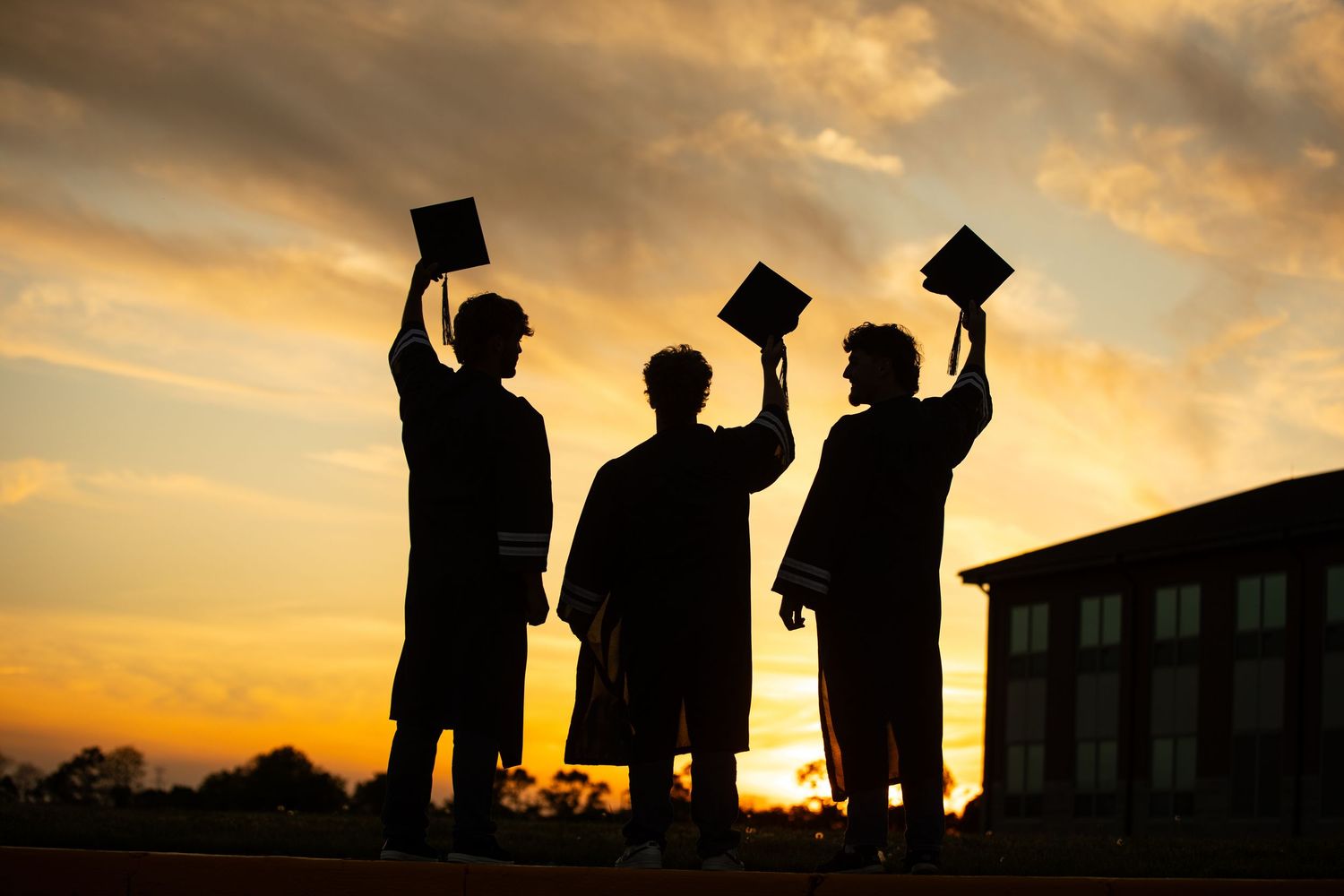 Three graduates in caps and gowns silhouetted against a golden sunset sky, raising their diplomas in celebration.